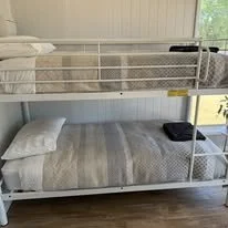 An indoor room with a bunk bed, featuring a mattress and pillows on the bottom bunk and a mattress with pillows on the top bunk, positioned near a window with blinds.