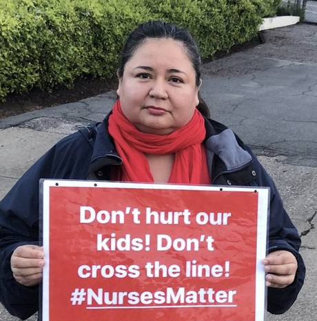 Woman holding a sign that reads, "Don't hurt our kids! Don't cross the line! #NursesMatter" outside near bushes and pavement.