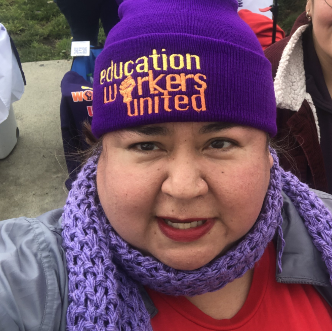 Close-up of a woman wearing a purple beanie with yellow and orange text, purple knitted scarf, and red lipstick, outdoors with people and trees in the background.