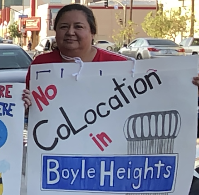 A woman holding a protest sign that reads 'No CoLocation in Boyle Heights' with a drawing of a lamppost.