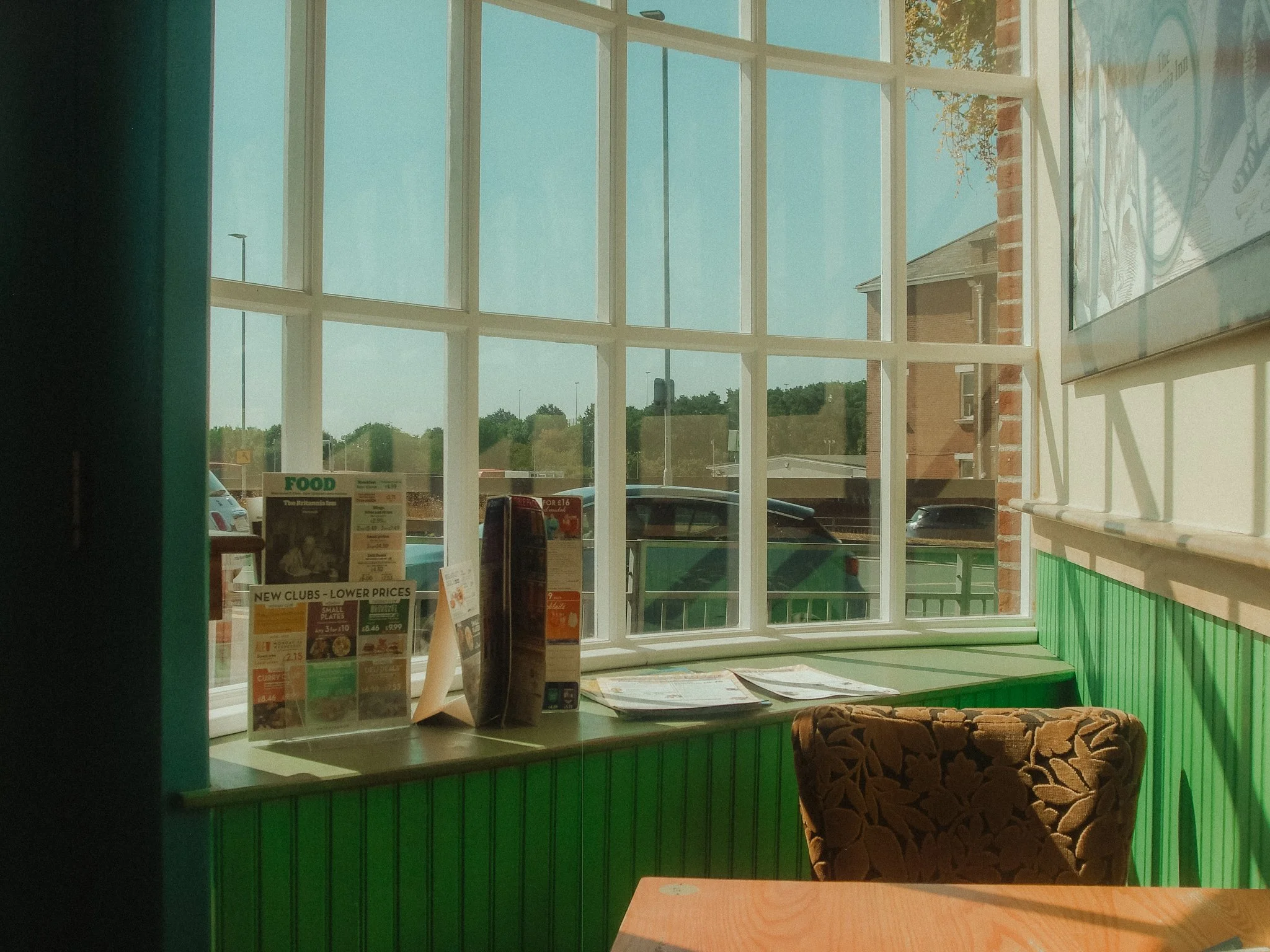 Inside a fast food restaurant or cafe, looking out a large window with green trim, with advertisements and a brown patterned chair visible.