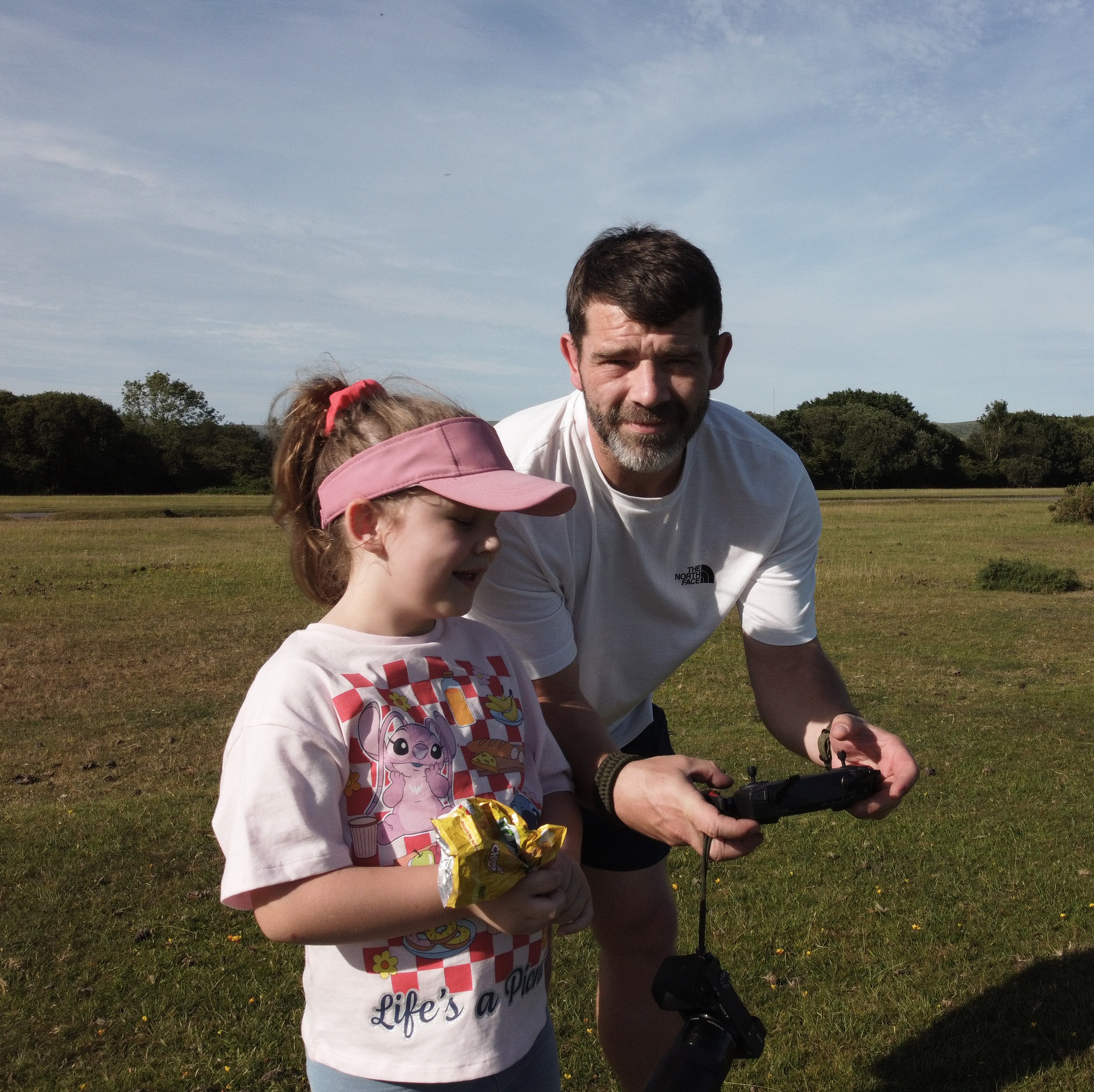 A man and a young girl outdoors in a grassy field, with the man holding a remote control and the girl looking at it, as they enjoy a sunny day.