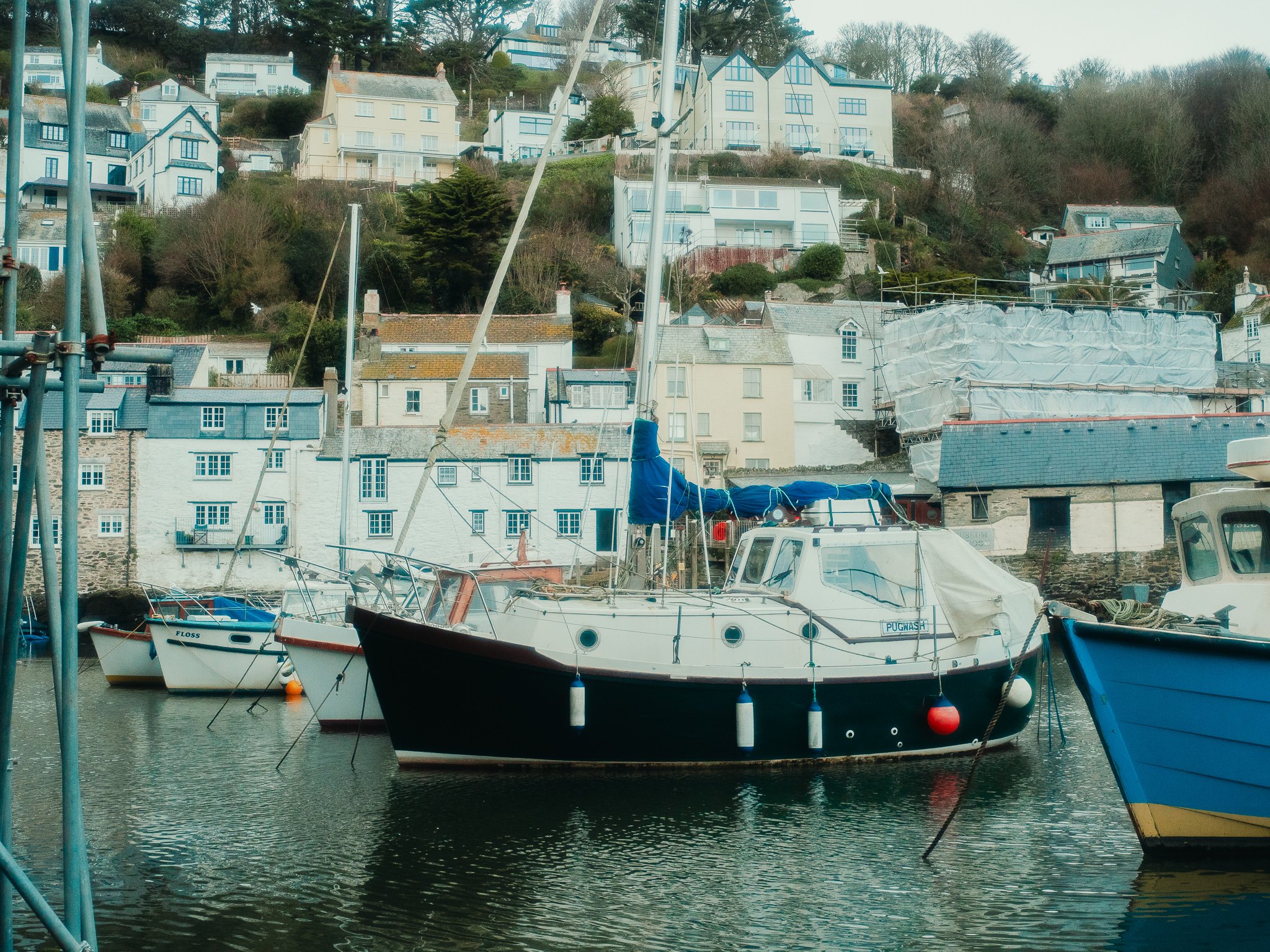 A small harbor with sailboats docked in the water and multi-story houses on a hillside in the background.