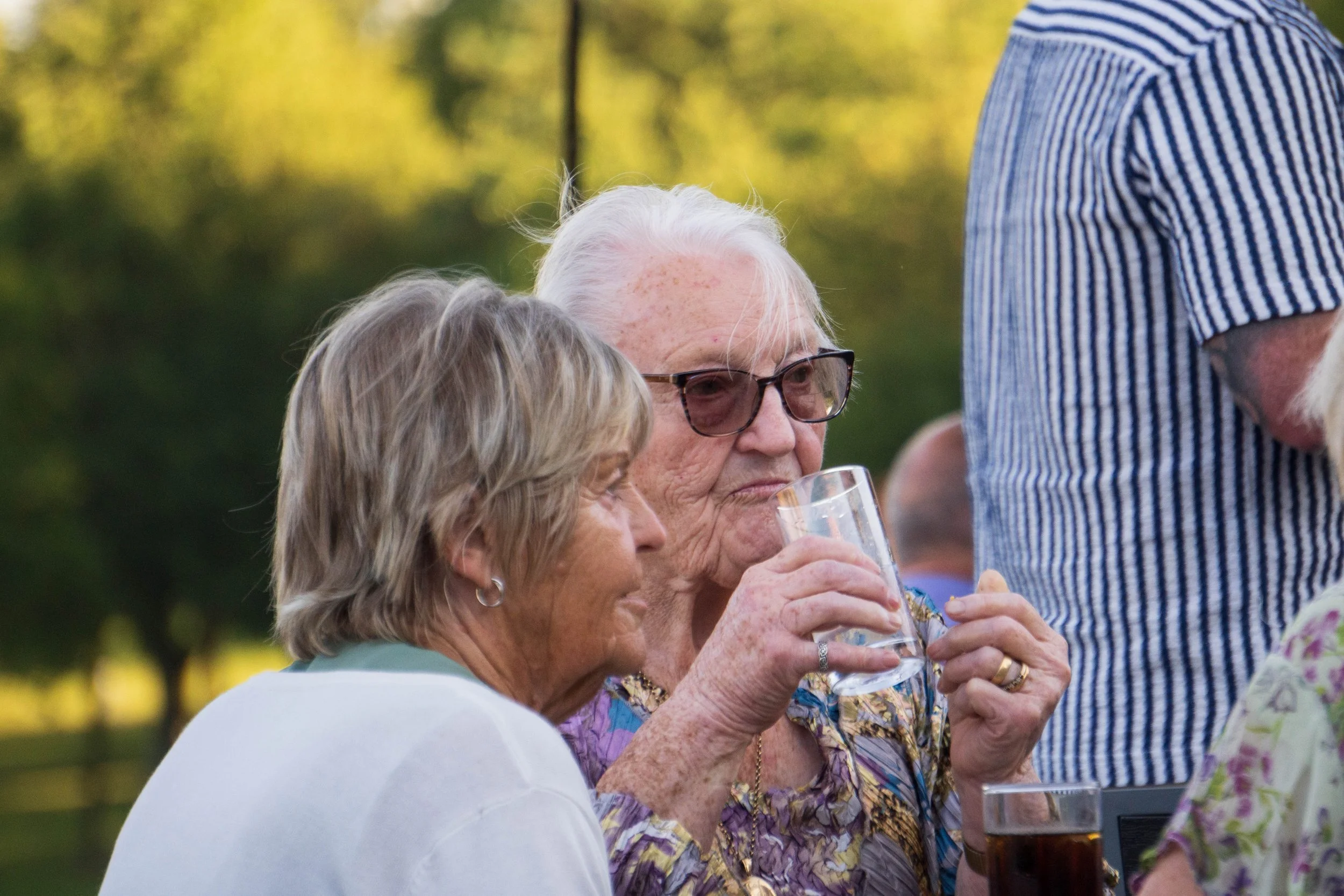 Older woman with white hair and glasses holding a glass of beverage, engaging in conversation with another woman outdoors.