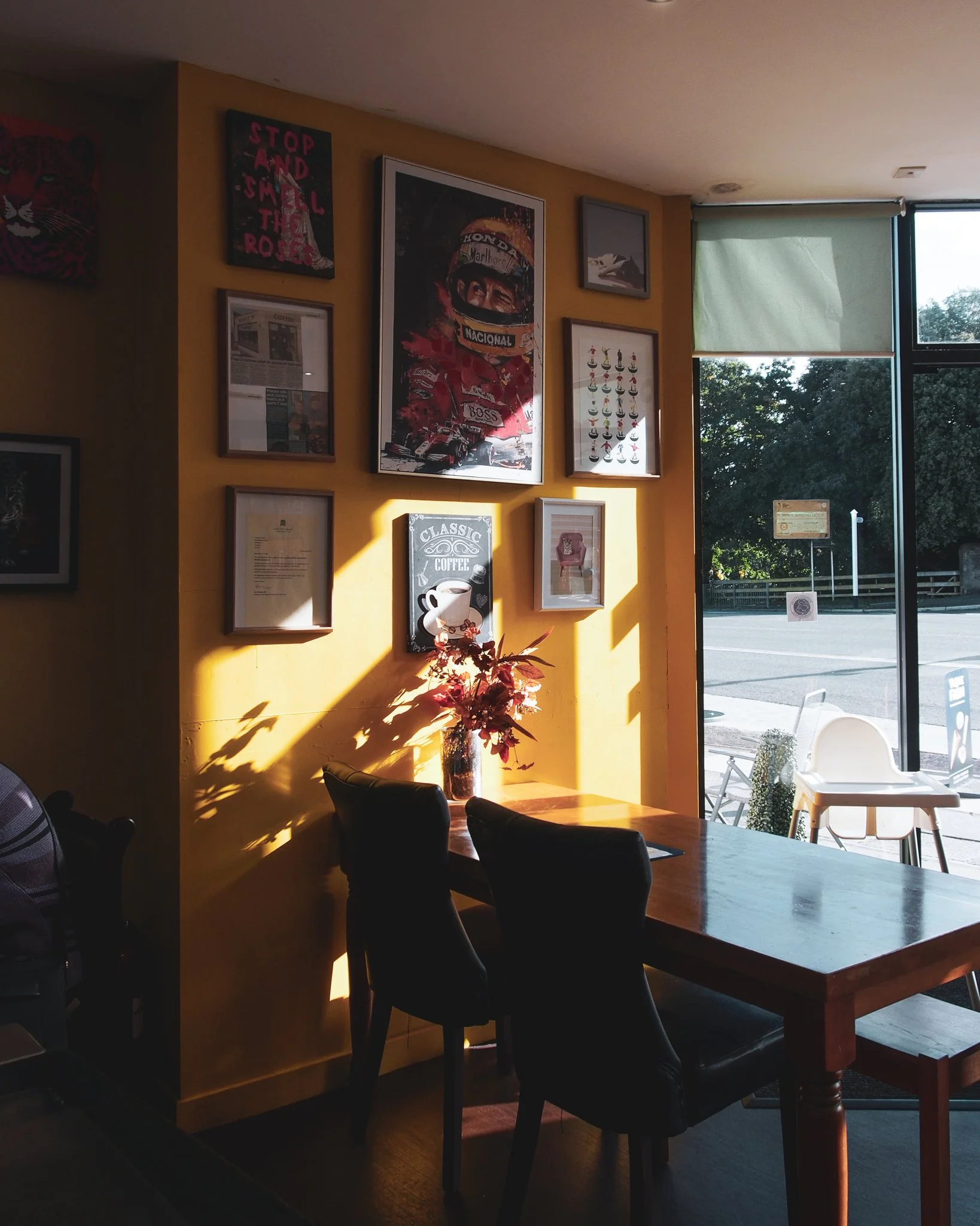 Sunlight casts shadows on a yellow wall decorated with framed pictures and posters inside a cafe. Chairs and a wooden table are nearby, with a small vase of flowers on the table. Large windows reveal an outdoor street scene.