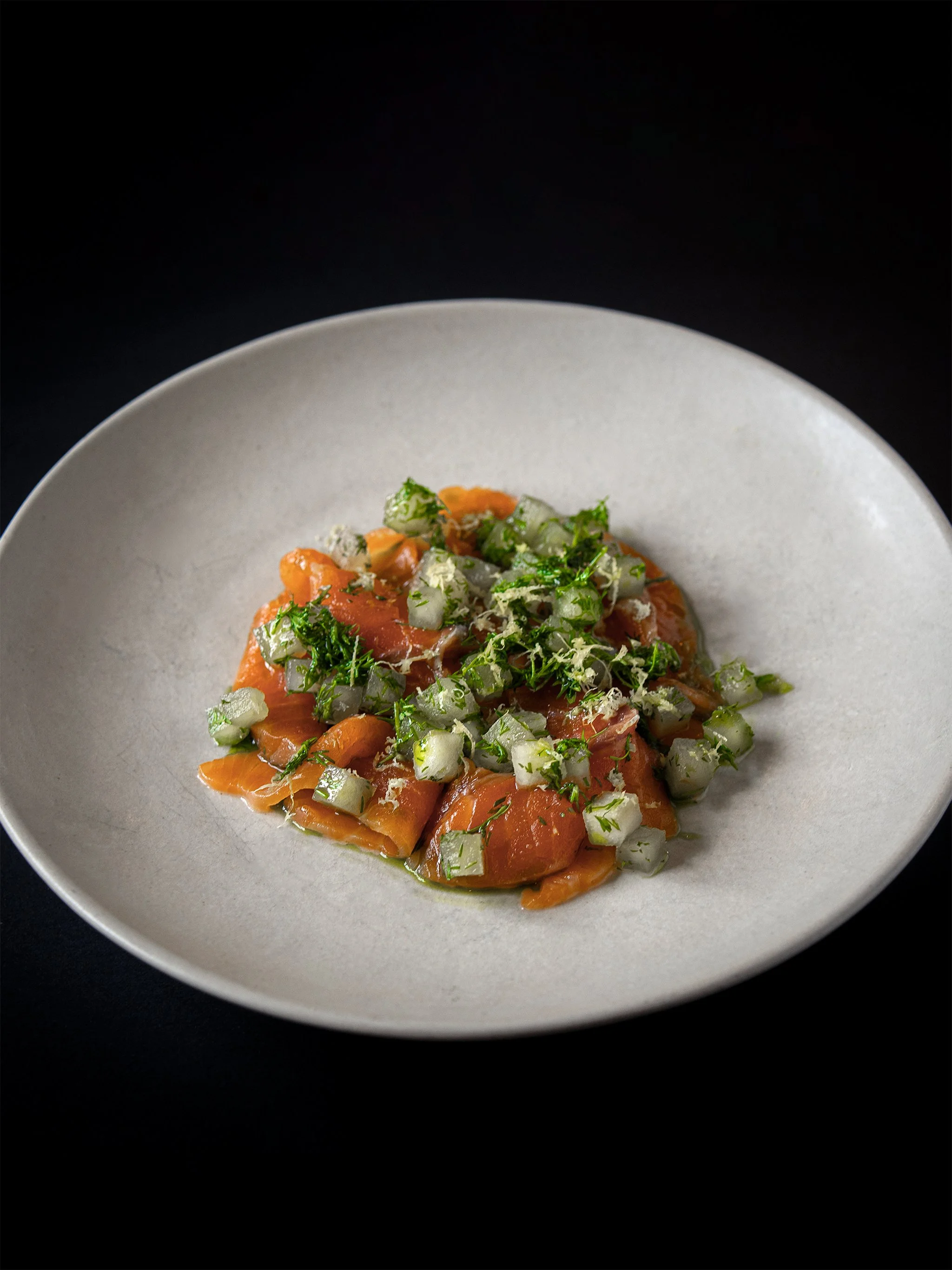 Plate of smoked salmon topped with chopped vegetables and herbs on a white dish against a black background.