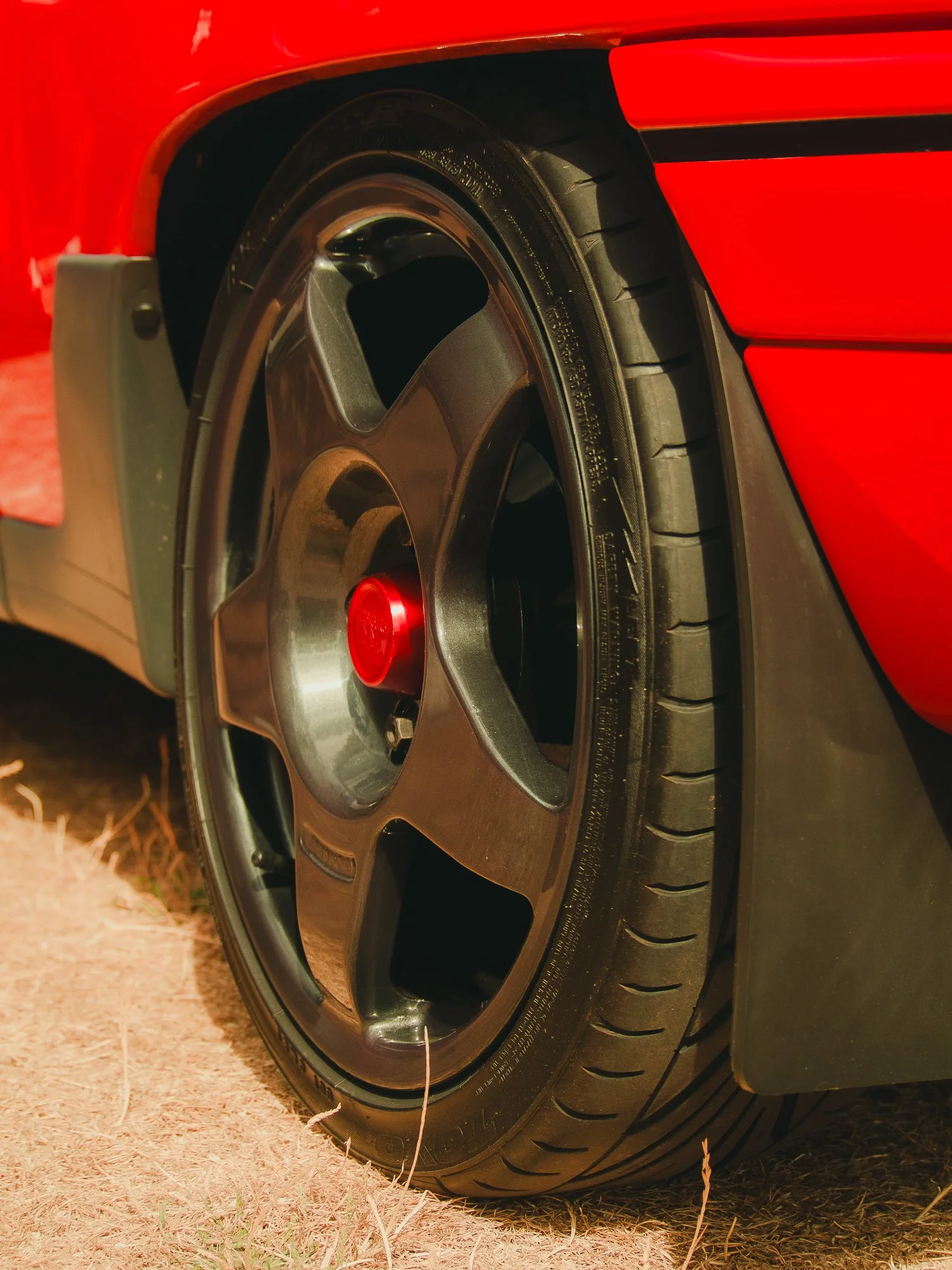 Close-up of a car tire and wheel on a red car, with part of the tire touching a dry grassy surface.