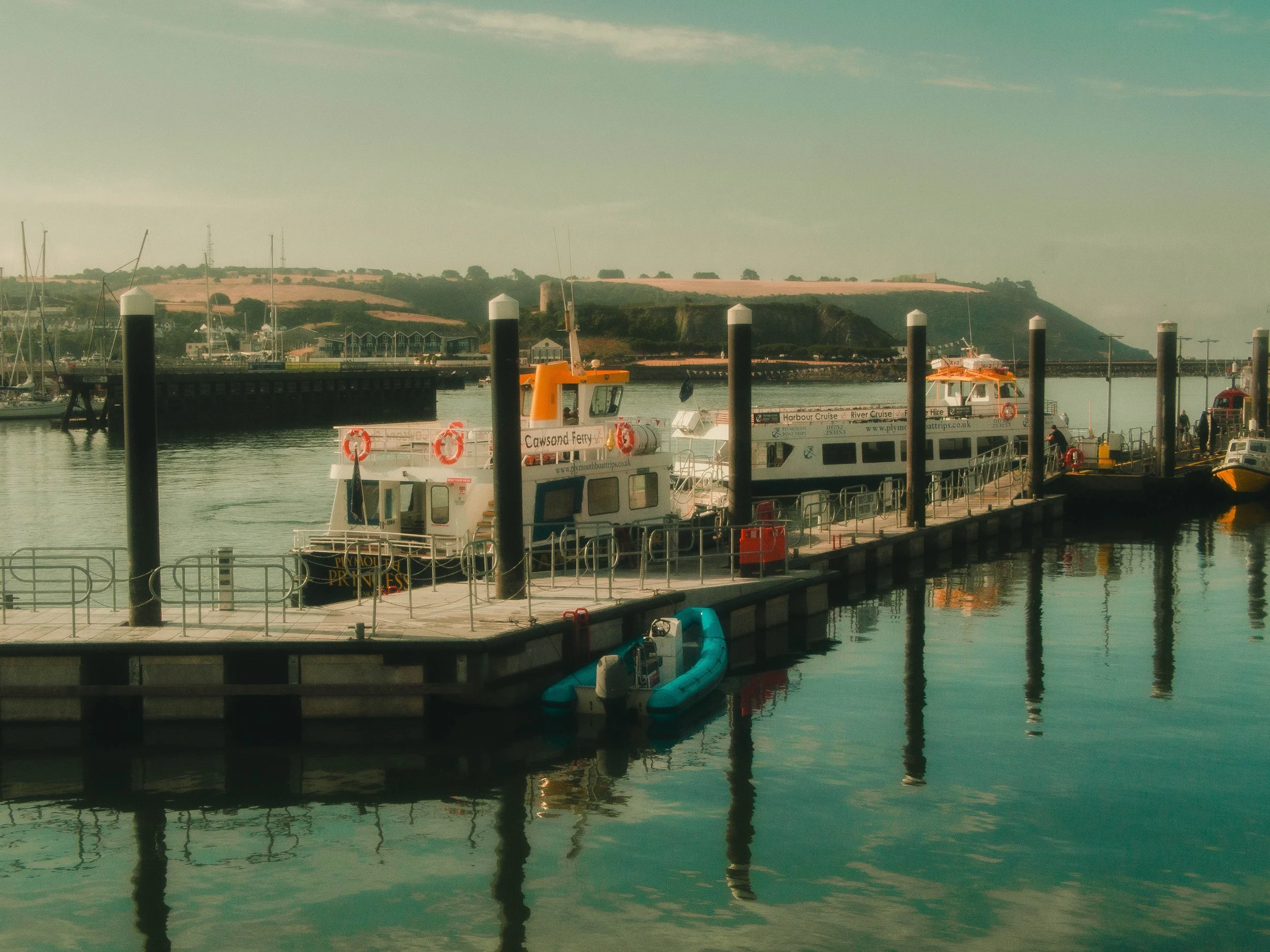 View of a harbor with boats docked, a ferry boat in the foreground, and a hilly landscape with buildings in the background.