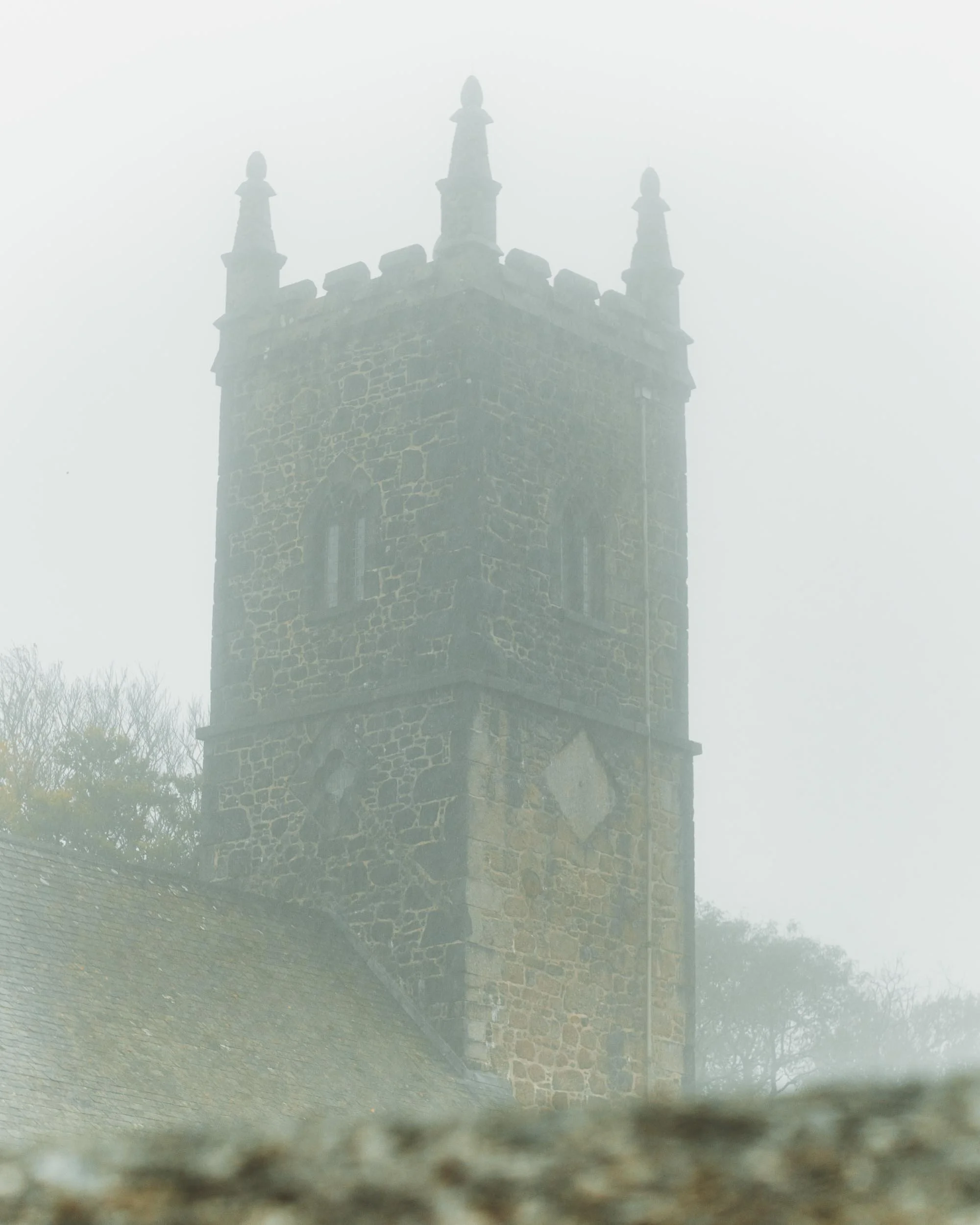 A foggy view of a tall stone castle tower with decorative battlements and spires on top.