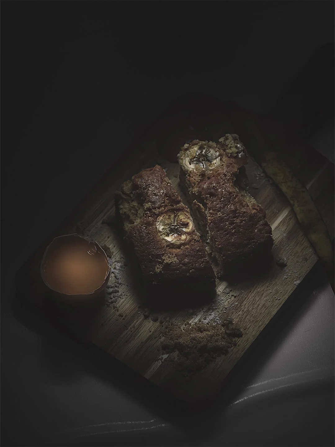 Two slices of banana bread on a wooden cutting board with a small cup of dipping sauce or syrup nearby.