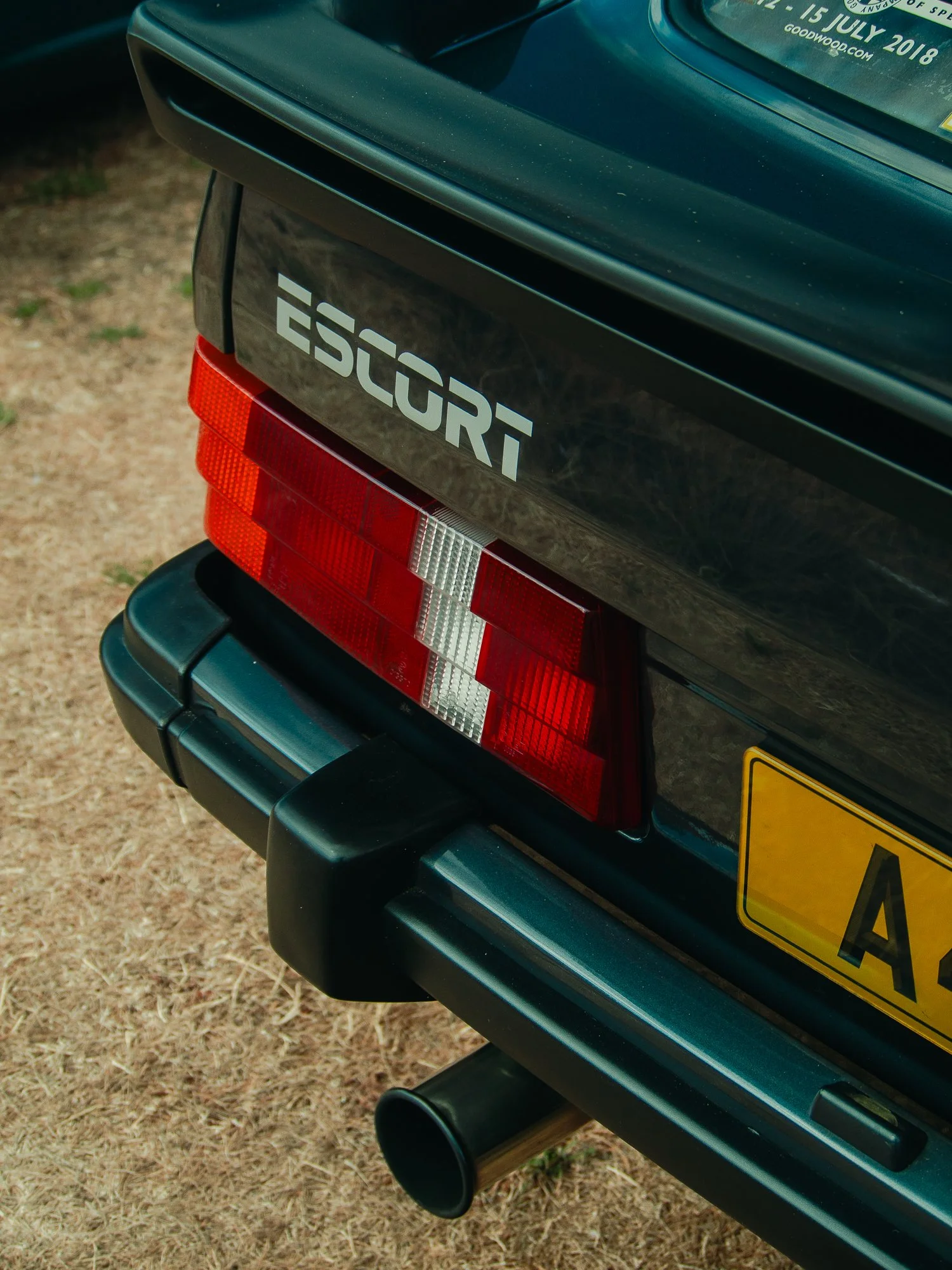 Close-up of the rear of a green Subaru car showing the taillight, exhaust pipe, part of the rear bumper, and a sticker on the rear window indicating the date of an event.