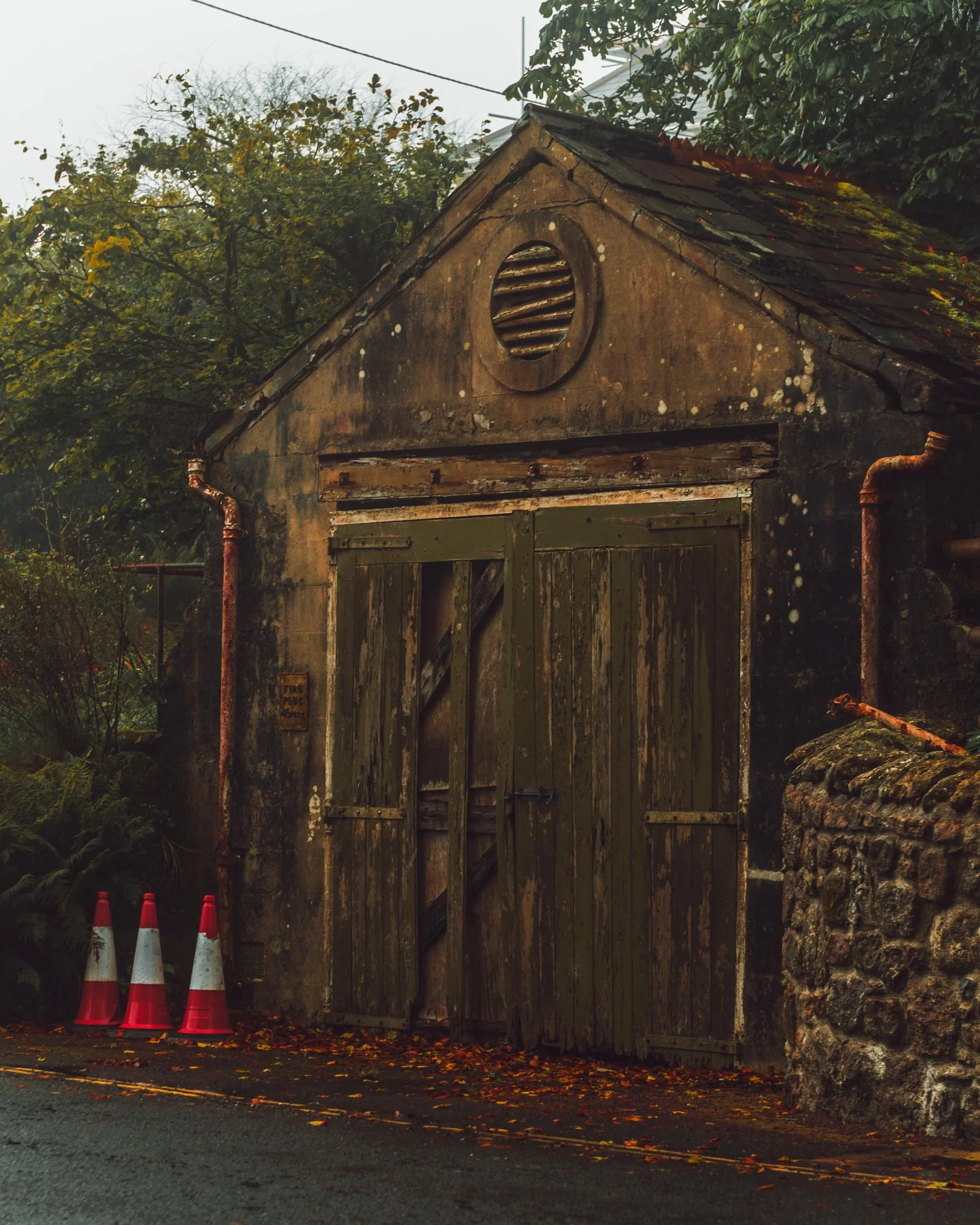 An old, weathered building with a boarded-up door and a circular vent near the peak of the roof, surrounded by trees with yellowing leaves and a stone wall on one side. Three orange and white construction cones are at the bottom of the image, with fa