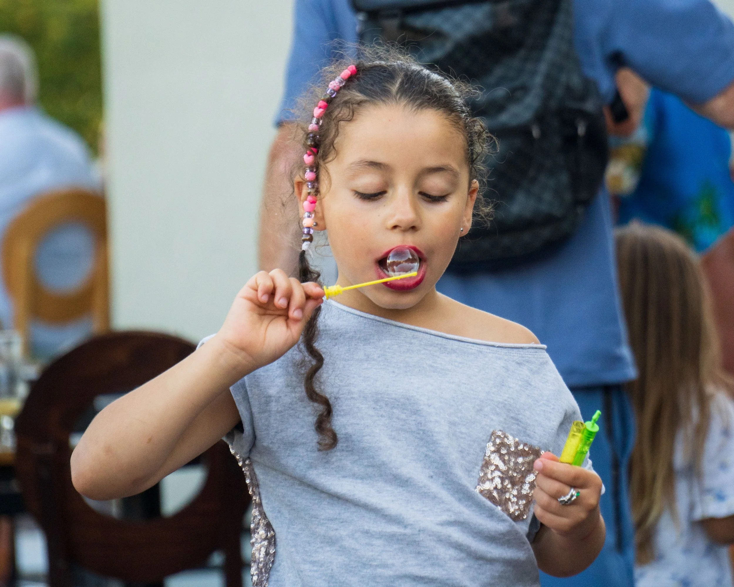 A young girl with curly hair, wearing a gray shirt with sequined sleeves, blowing bubbles with a yellow bubble wand during an outdoor gathering.