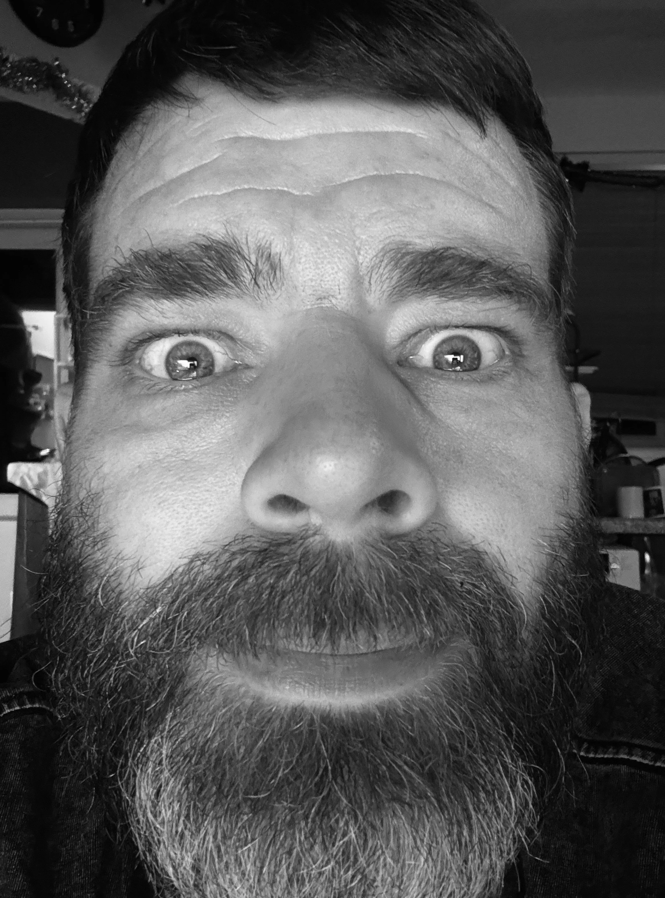 Close-up of a man's face with a beard and mustache, wide-open eyes, and a surprised or playful expression, taken in black and white.