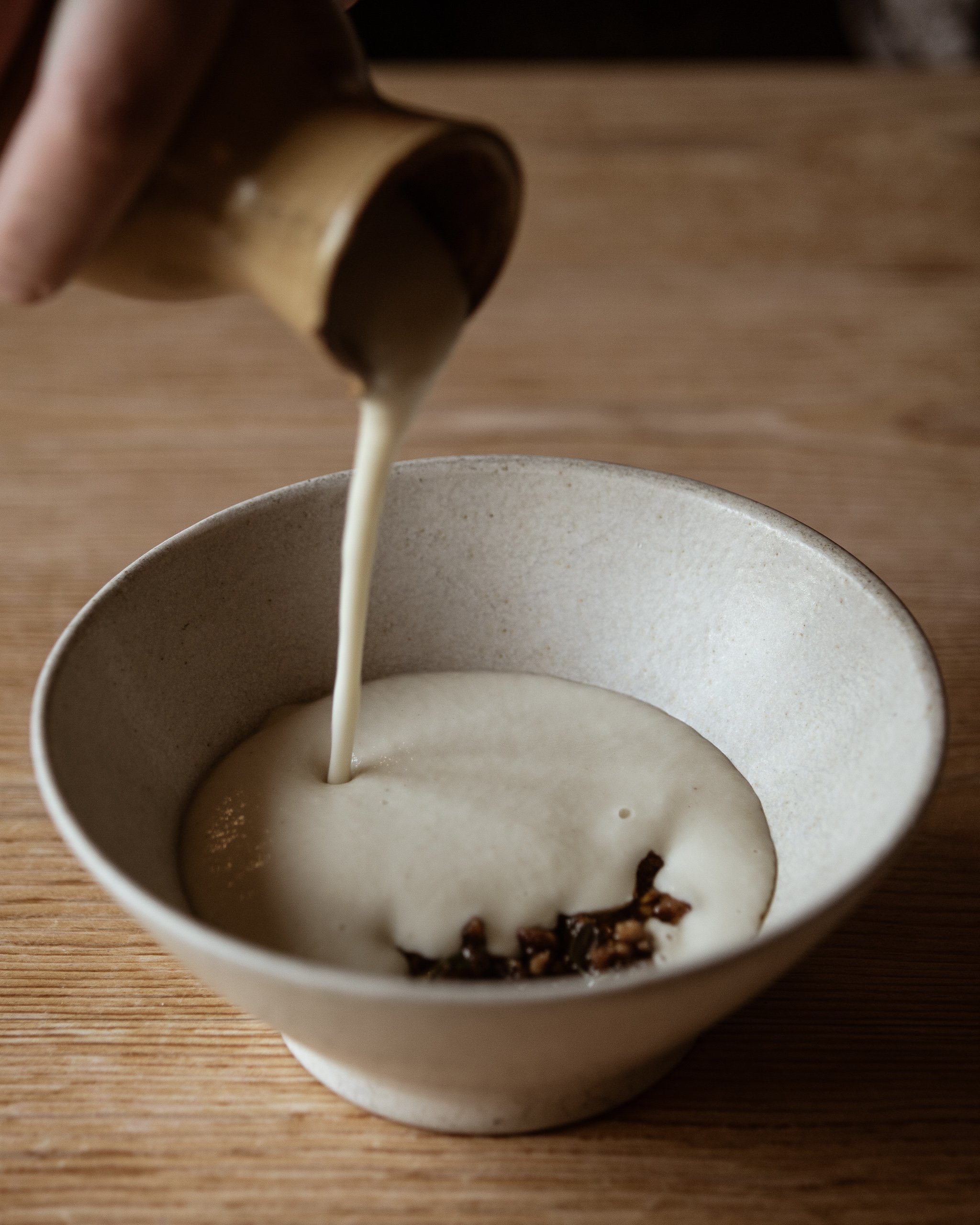 Cream being poured from a small brown bottle into a white bowl with some dark pieces in it, on a wooden surface.