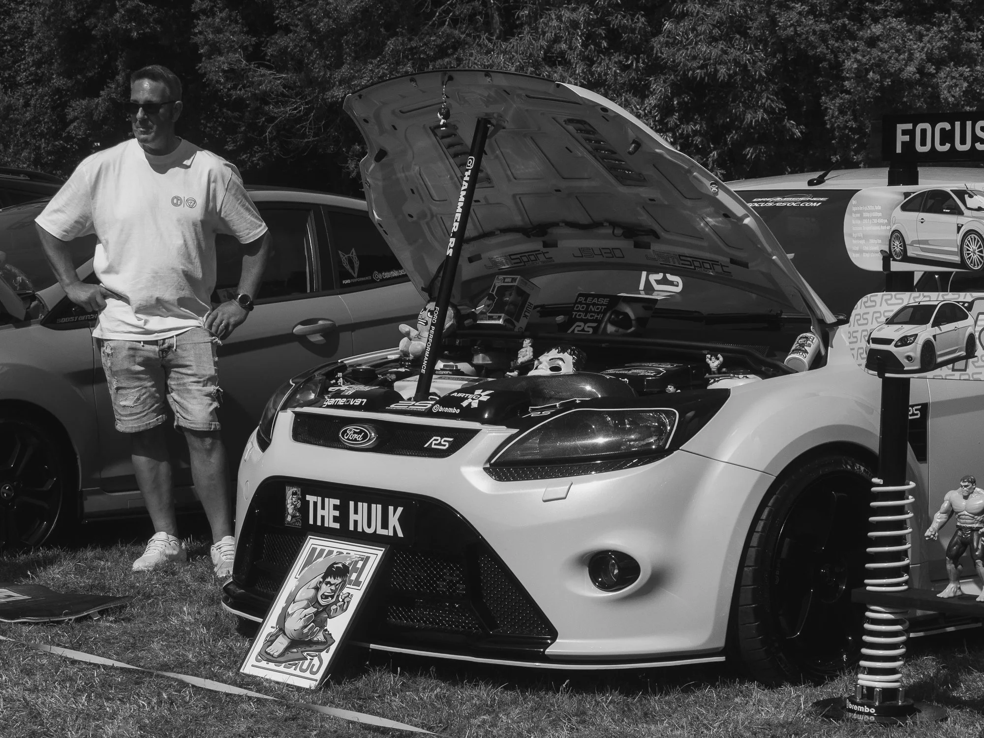 A man wearing sunglasses, a white T-shirt, and shorts stands beside a white Ford Mustang with its hood open at a car show. The car features a graphic of the Hulk on the front grille. Behind, there are other cars and car-themed banners.