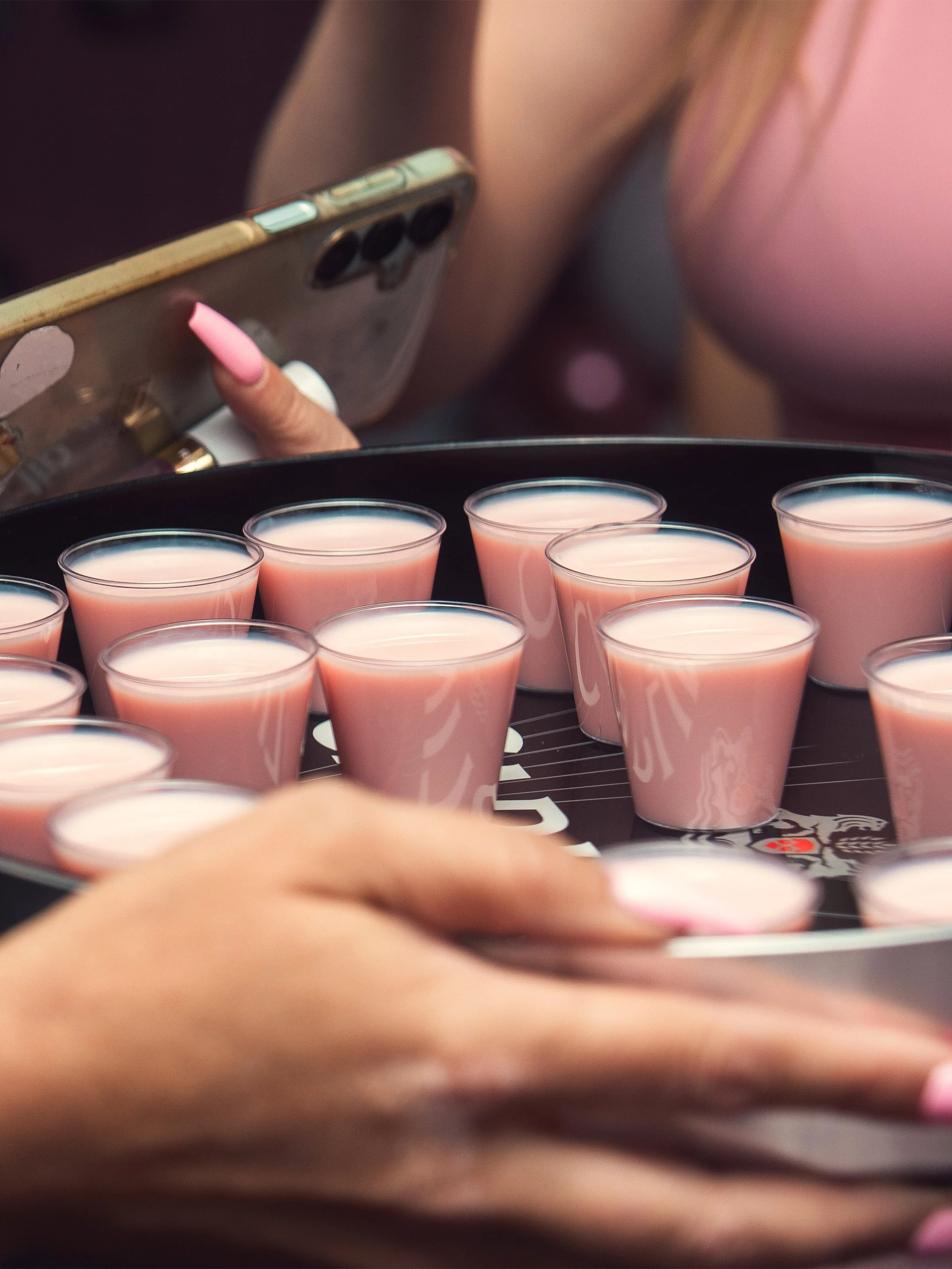 Close-up of a person holding a smartphone with a multicolor case, taking a photo of several pink drinks in small plastic cups on a tray.
