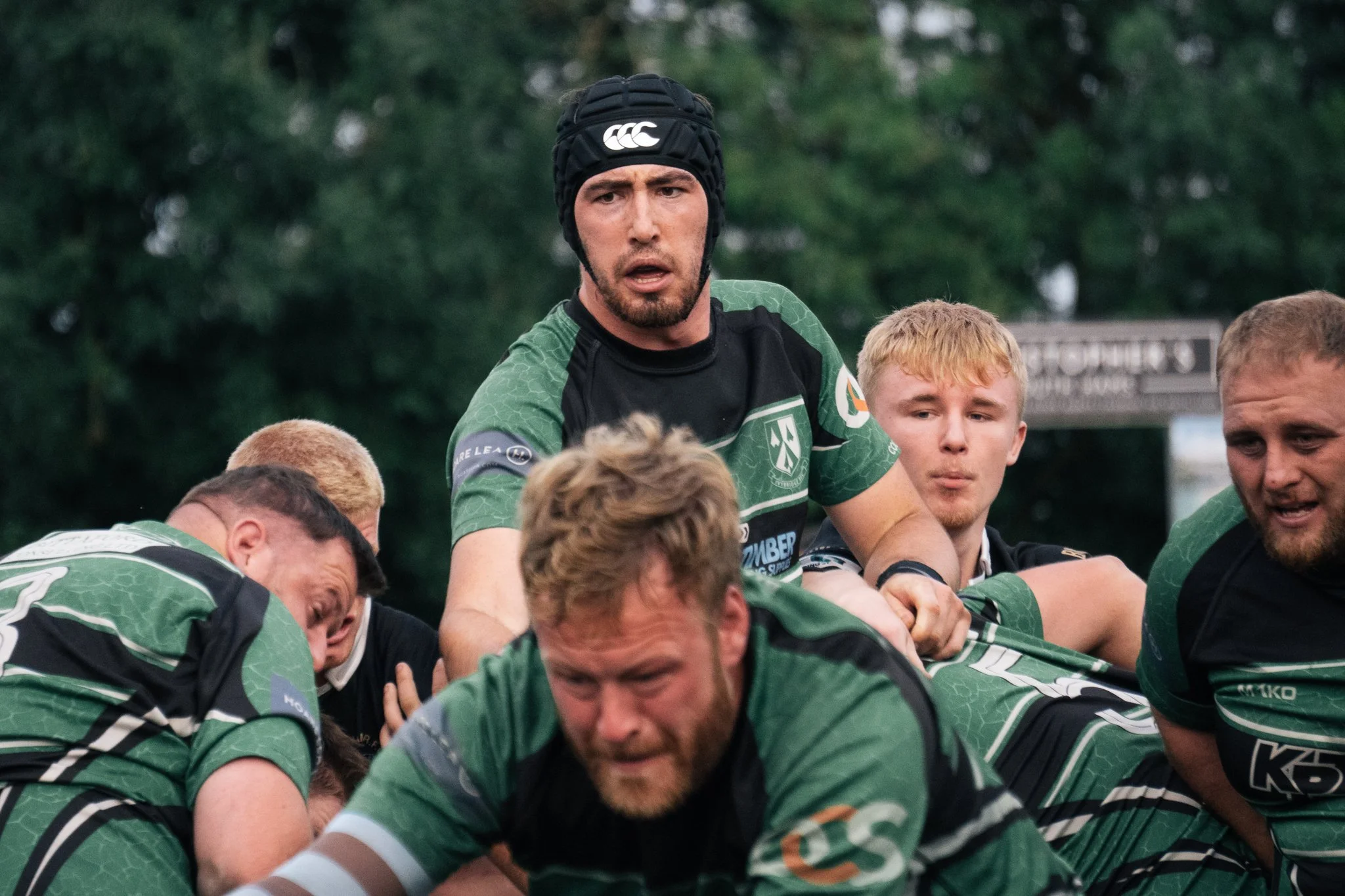 Rugby players in green and black uniforms engaging in a team huddle, with one player wearing a black protective headgear, during an outdoor rugby game.