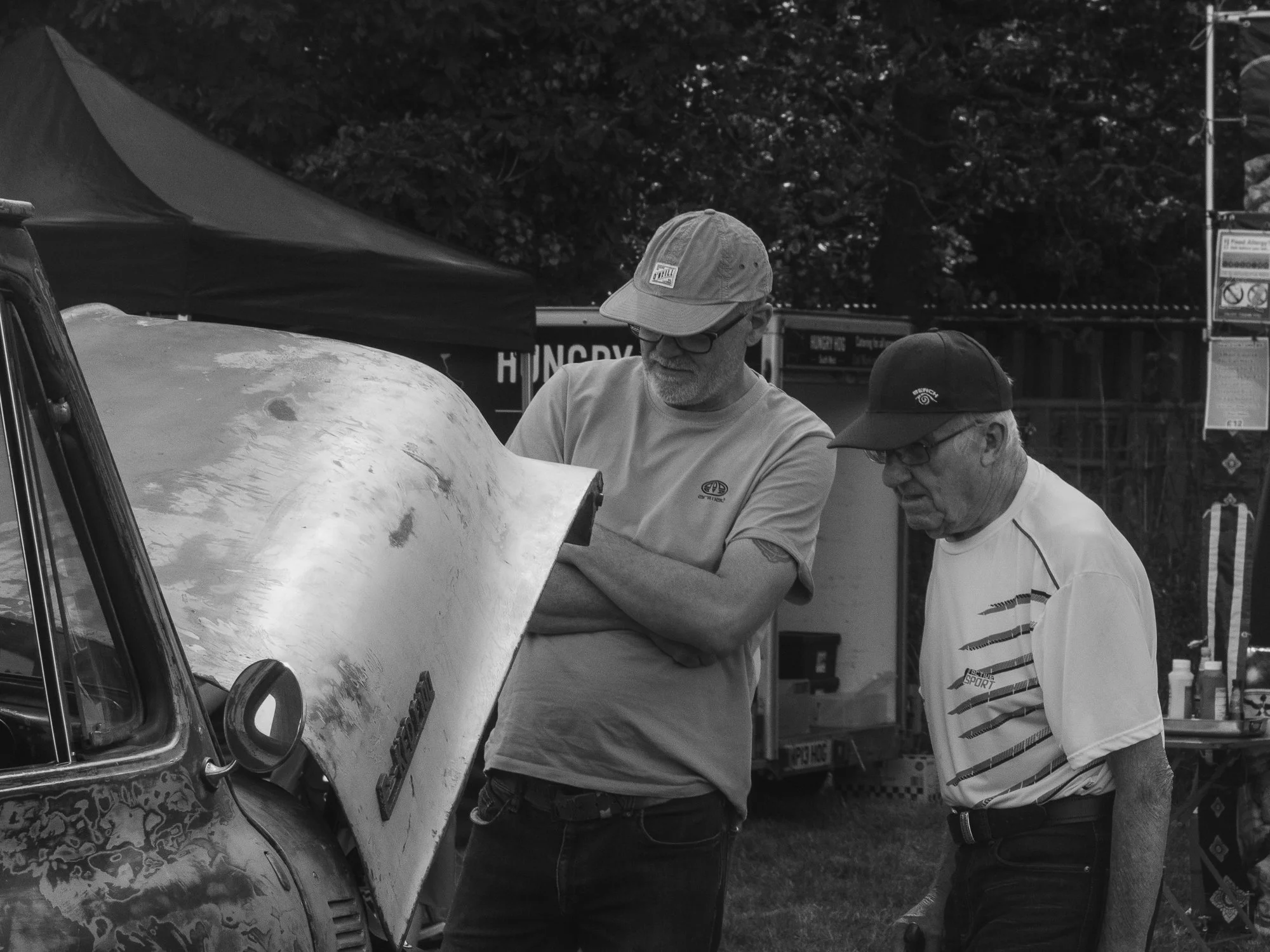 Two men are examining a vintage car at an outdoor event. One man with a cap and glasses is pointing at the car while the other man in a striped shirt looks on. There is a tent and trees in the background.