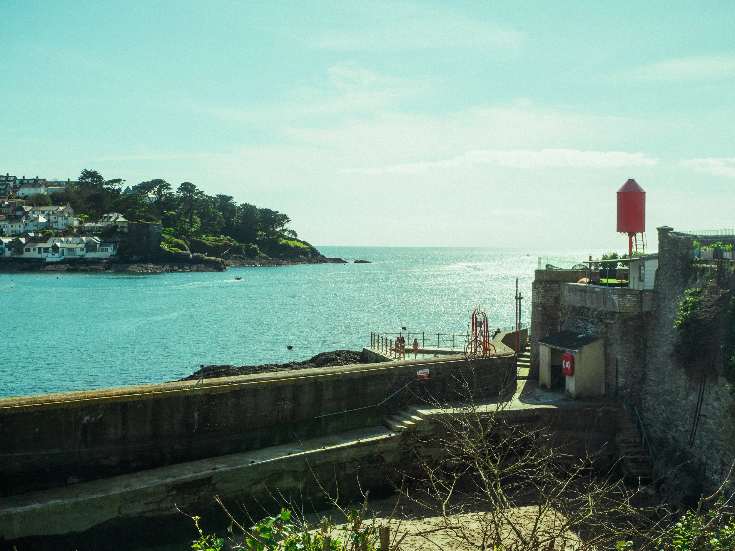 A coastal scene with houses on a hill to the left, a body of water in the foreground, a red lighthouse on the right, and a few people near the water.