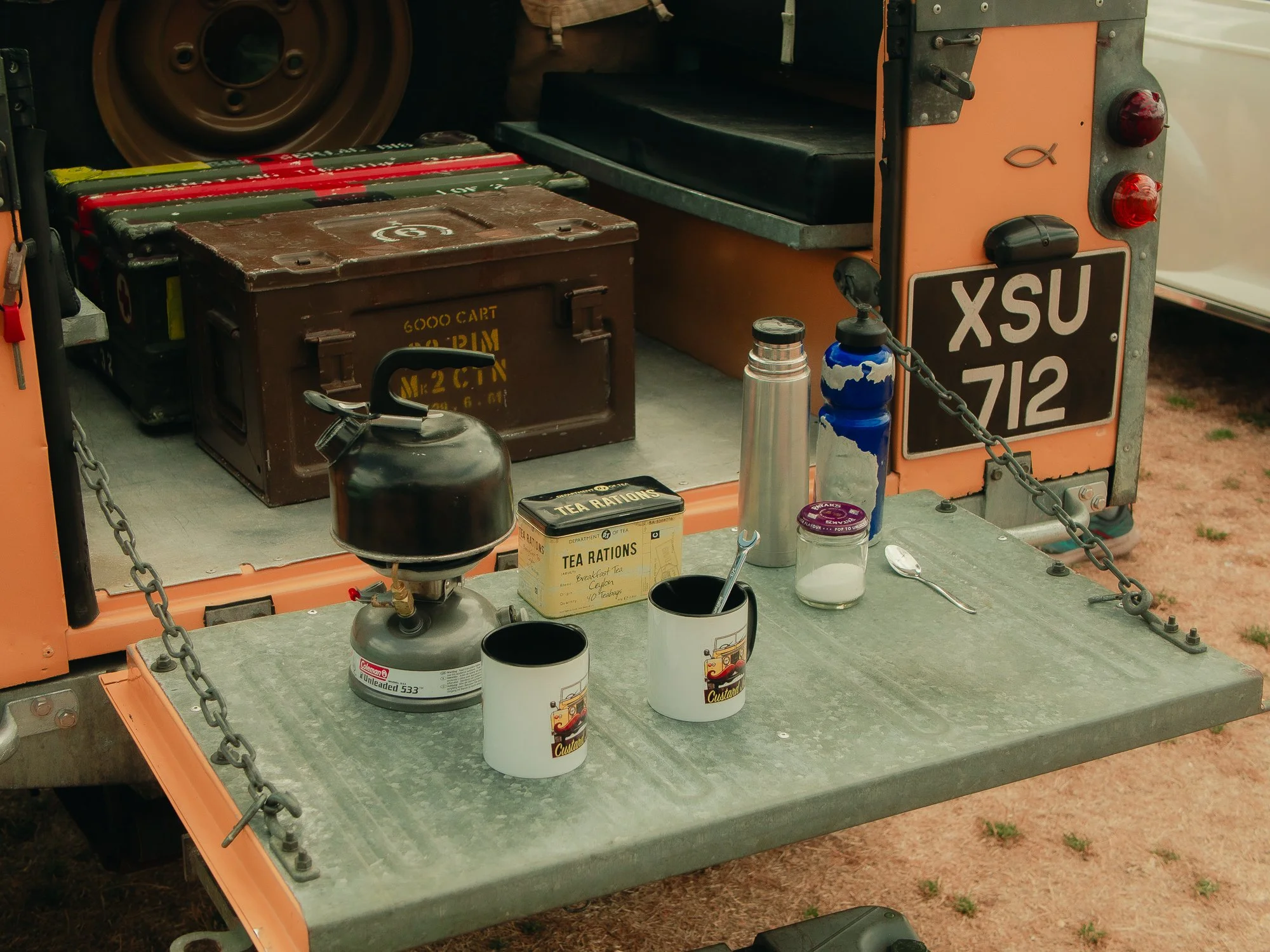 A vehicle back area with military-style ammunition boxes, coffee mugs, water bottles, and containers on a metal surface.