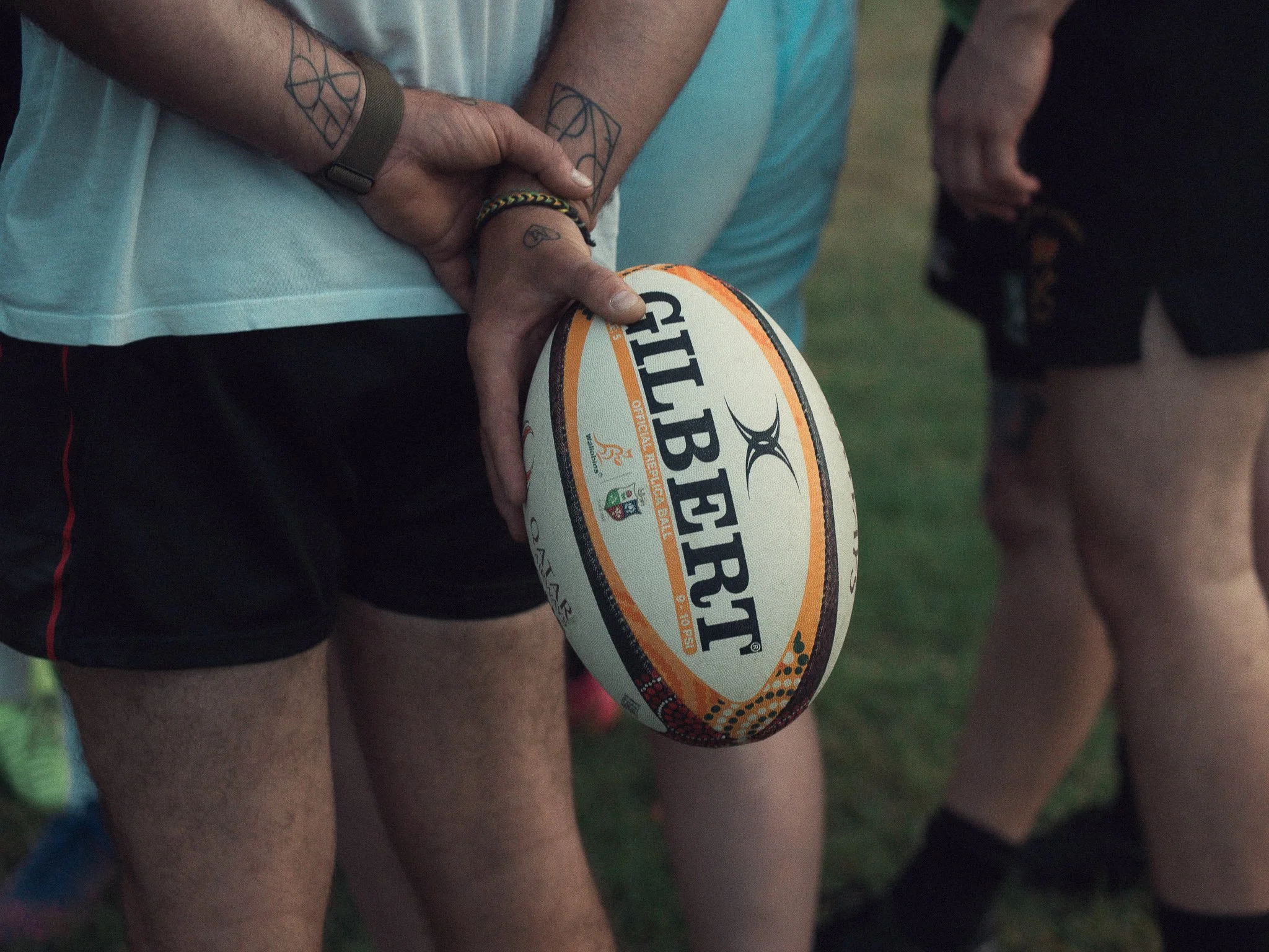 Person holding a rugby ball with the brand name "GILBERT" printed on it, standing outdoors with other people in the background.