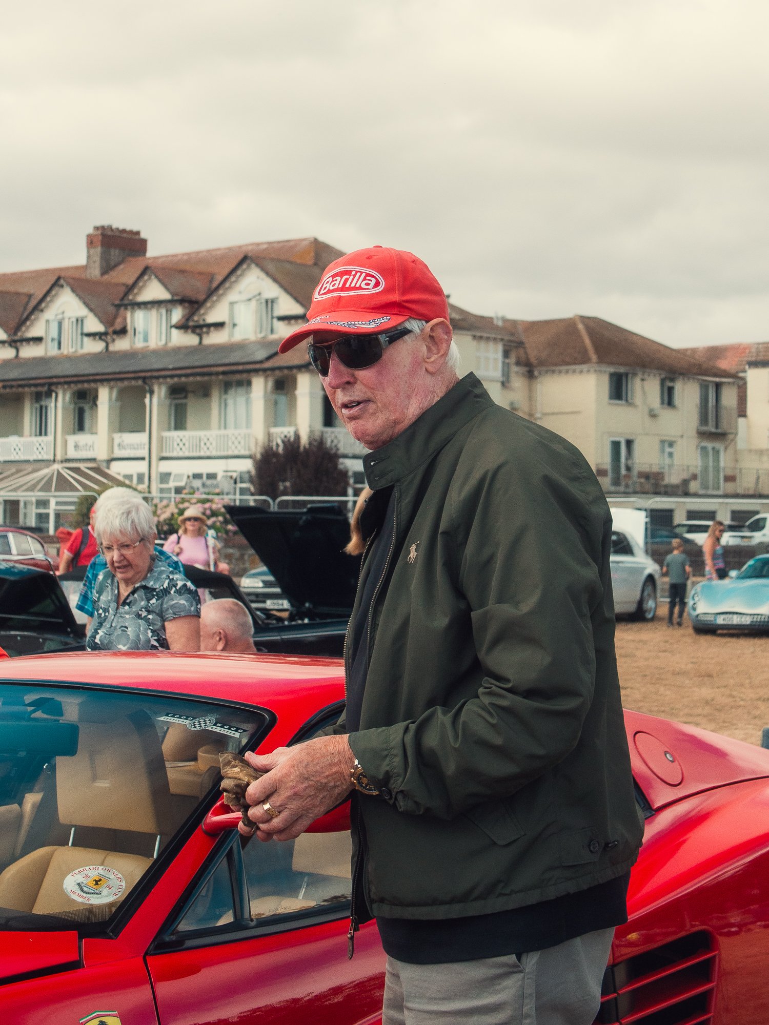 A man wearing sunglasses and a red Cap with 'Barilla' written on it, standing next to a red sports car at a car show. Several people and houses are in the background.