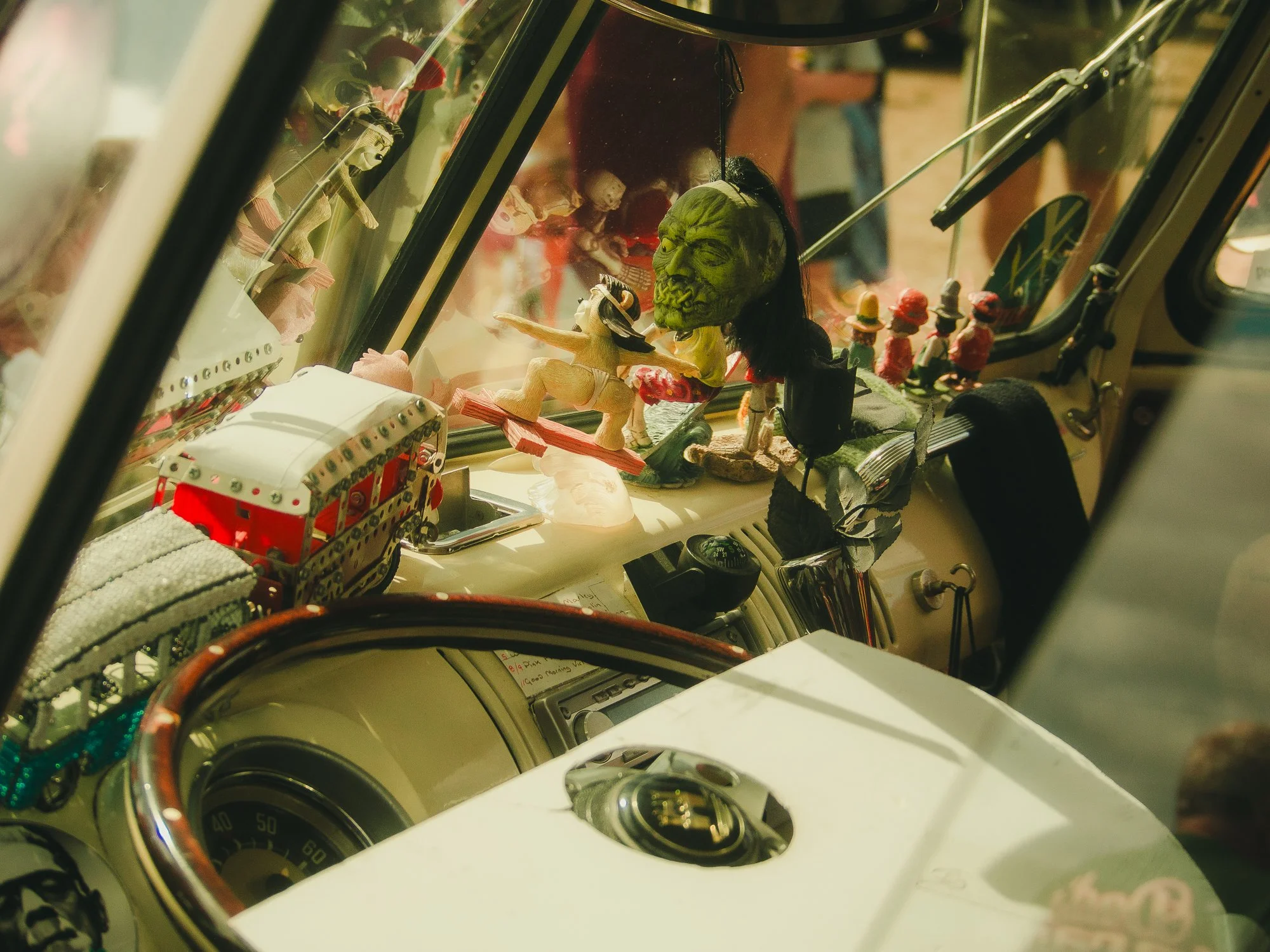 The image shows the interior of a vehicle with a collection of figurines including a green face monster, a reclining nude woman, and various tiny figures displayed behind a glass cover.