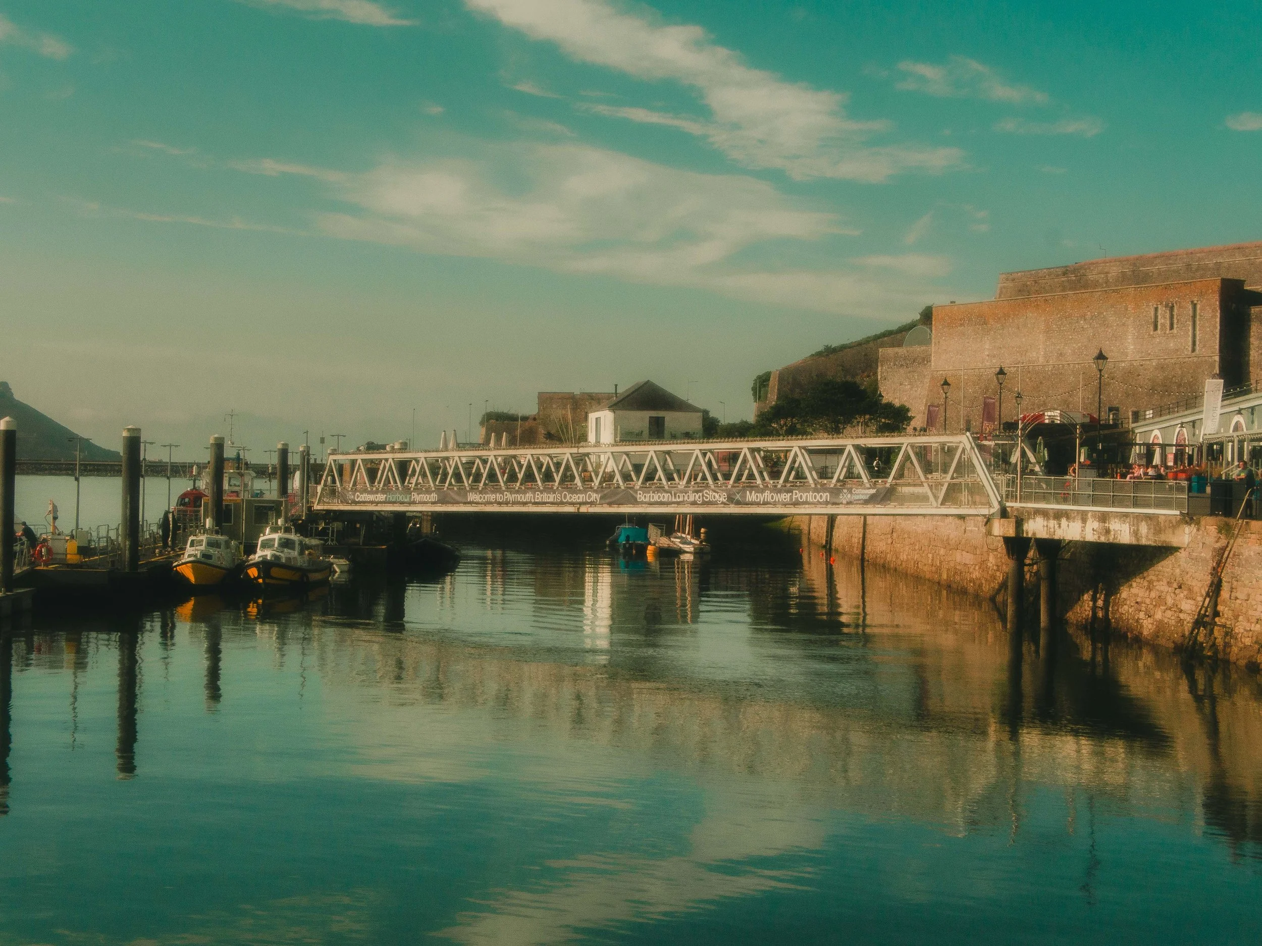 A waterfront scene at Cattewater Harbour in Plymouth, Britain. There are boats docked in the harbor, historic stone buildings on a hill, and a clear sky with some clouds.