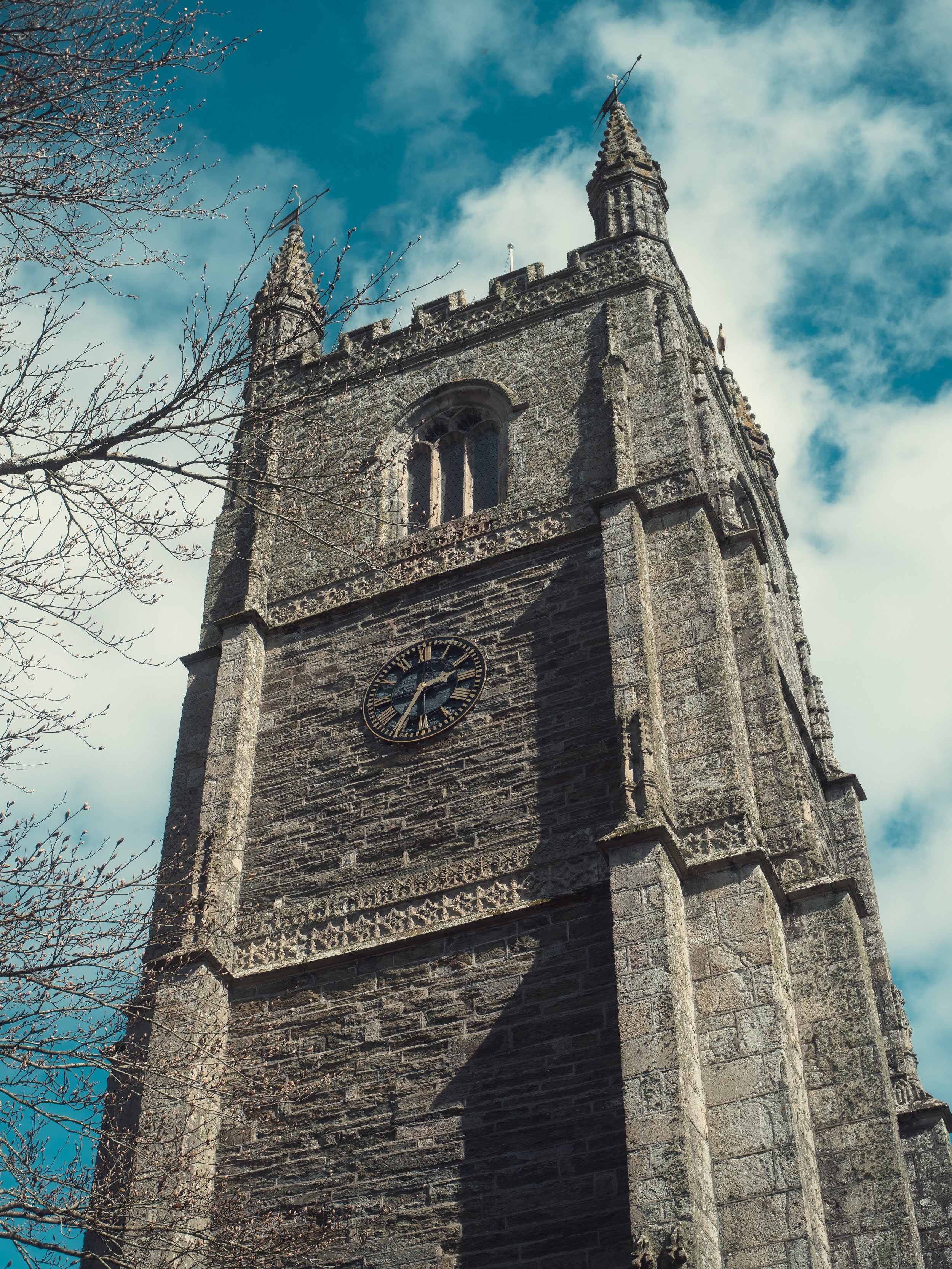 A historic stone church with a clock face and two tall steeples topped with crosses, under a partly cloudy sky, with leafless tree branches in the foreground.