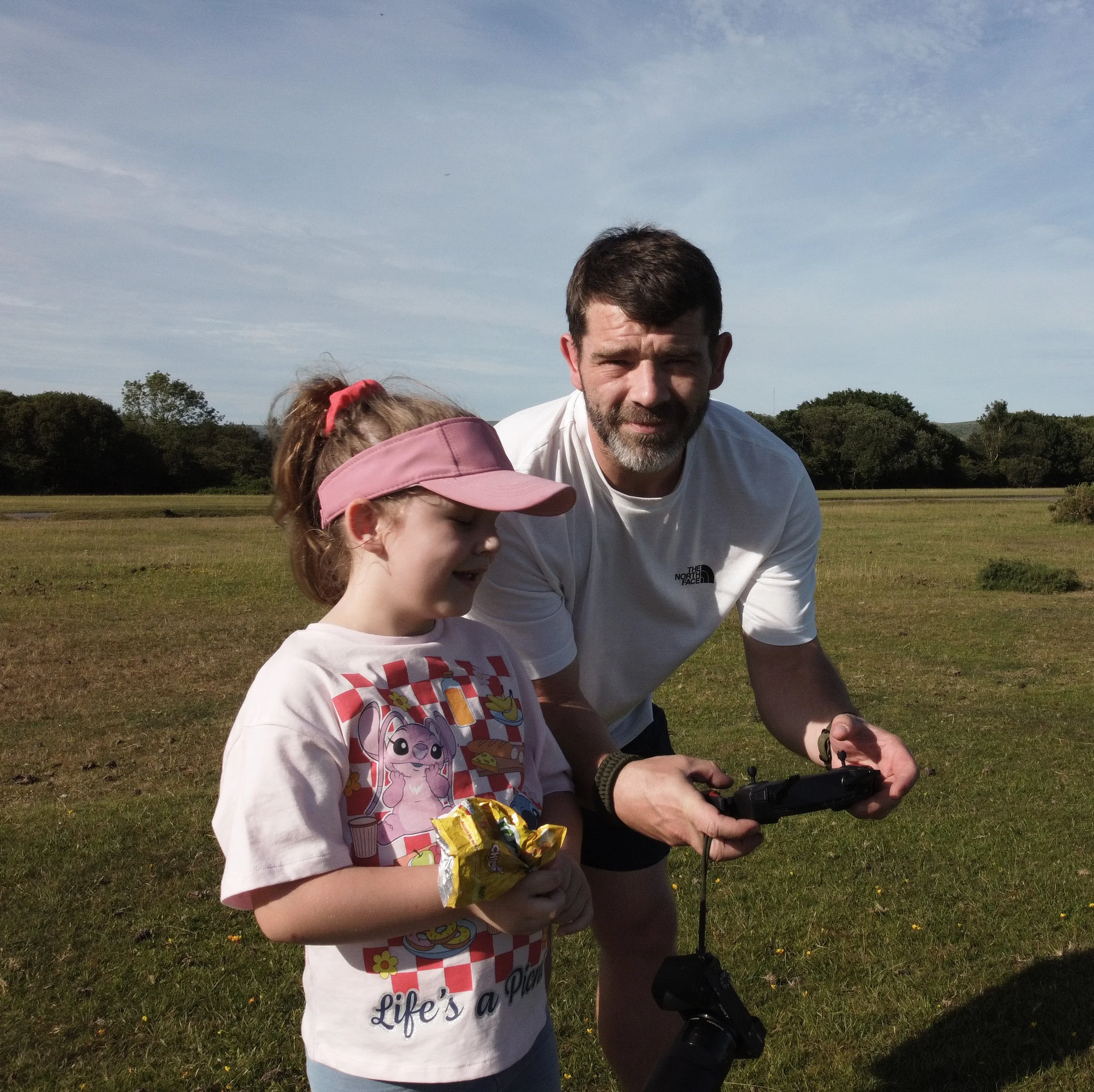 A young girl in a pink cap and a white T-shirt with a colorful graphic, standing outdoors on grass with a man, likely her father, crouching next to her. The man has dark hair and a beard, and is wearing a white T-shirt. They are in a park with trees and a cloudy sky in the background, holding a remote control and a bag of chips.