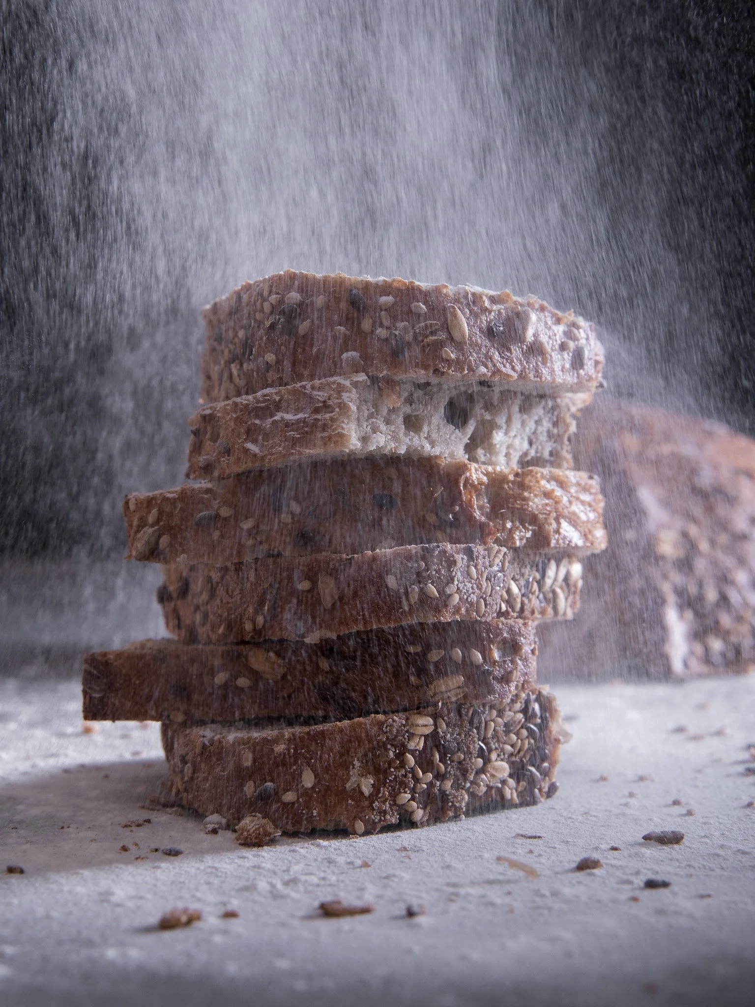 Stacked slices of multigrain bread with seeds, dusted with flour, under a fine spray of water.
