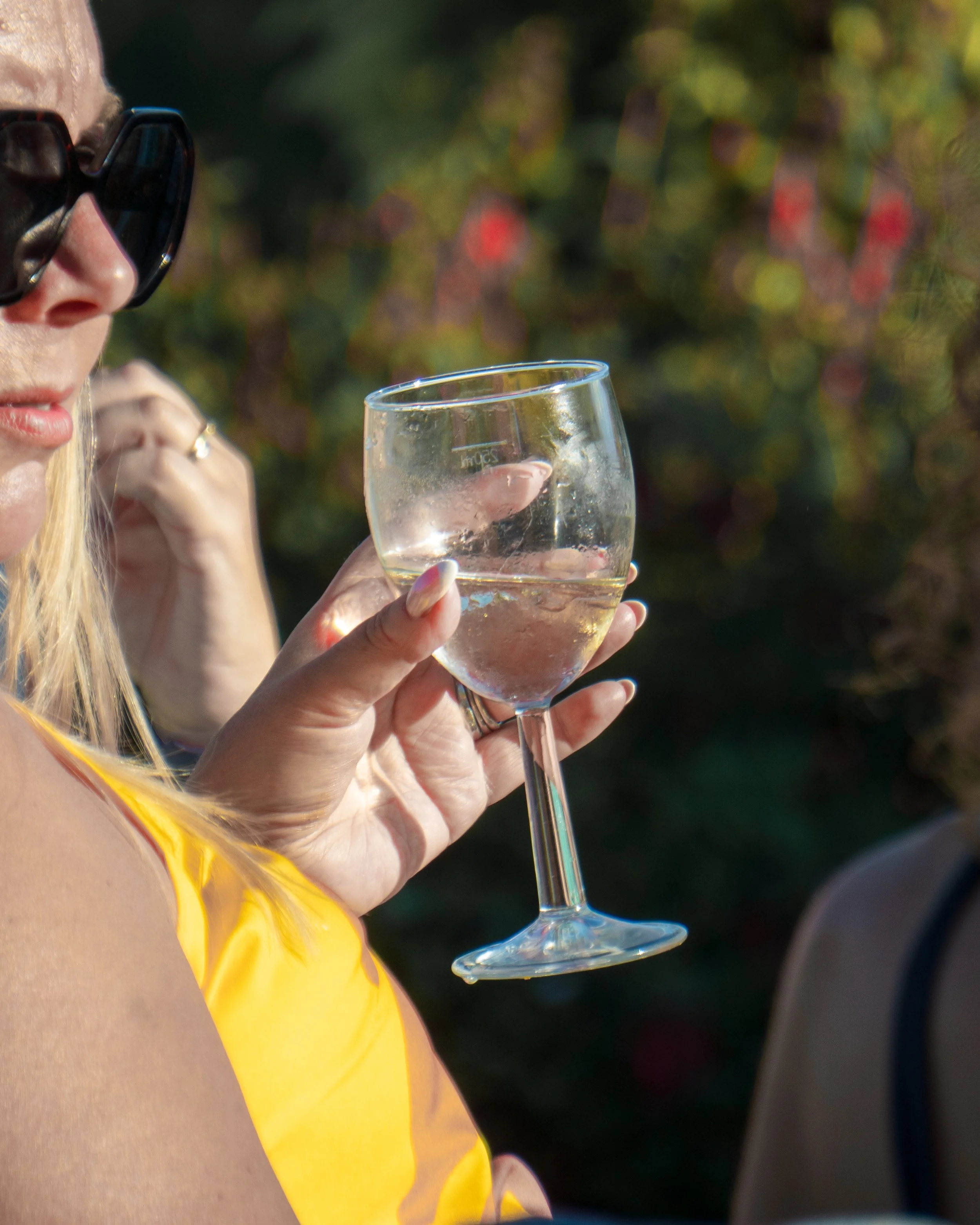 A woman in a yellow dress with blonde hair wearing black sunglasses holding a glass of white wine outdoors.