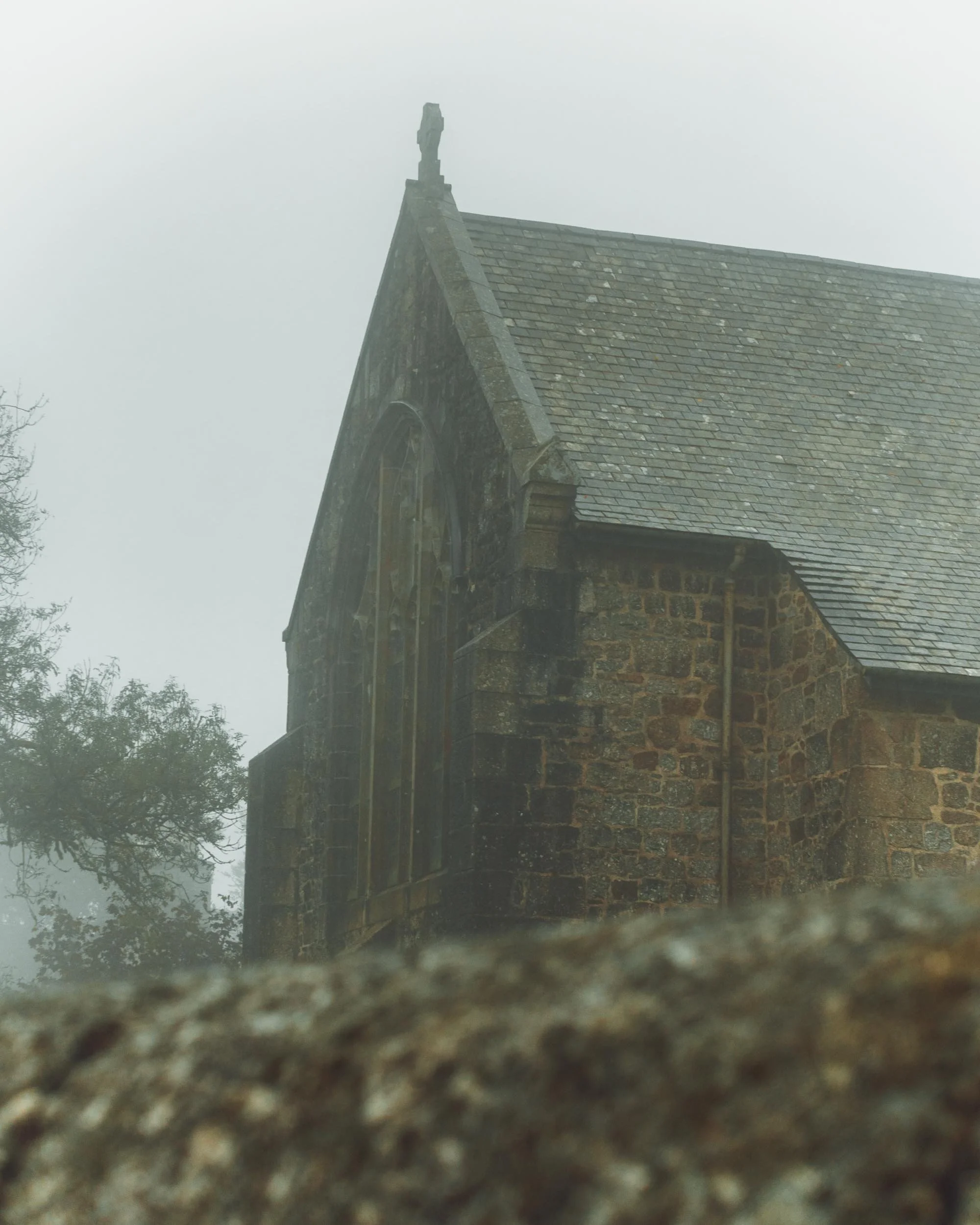 A stone church or chapel with a sloped roof and a small cross at the top of the gable end, covered in fog or mist.