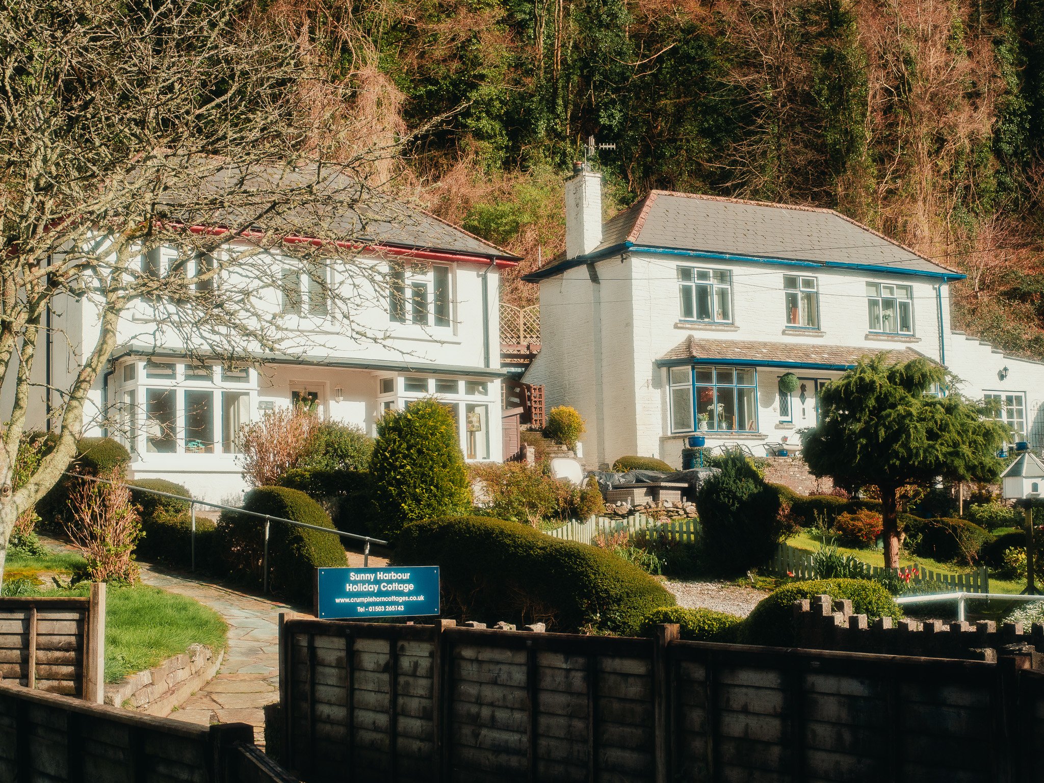 A large white house with a sloped roof, chimney, and multiple windows, surrounded by a well-maintained garden with various bushes and trees, with a blue sign in front that reads 'Sunny Harbour Holiday Cottage' and some small buildings or cottages in 