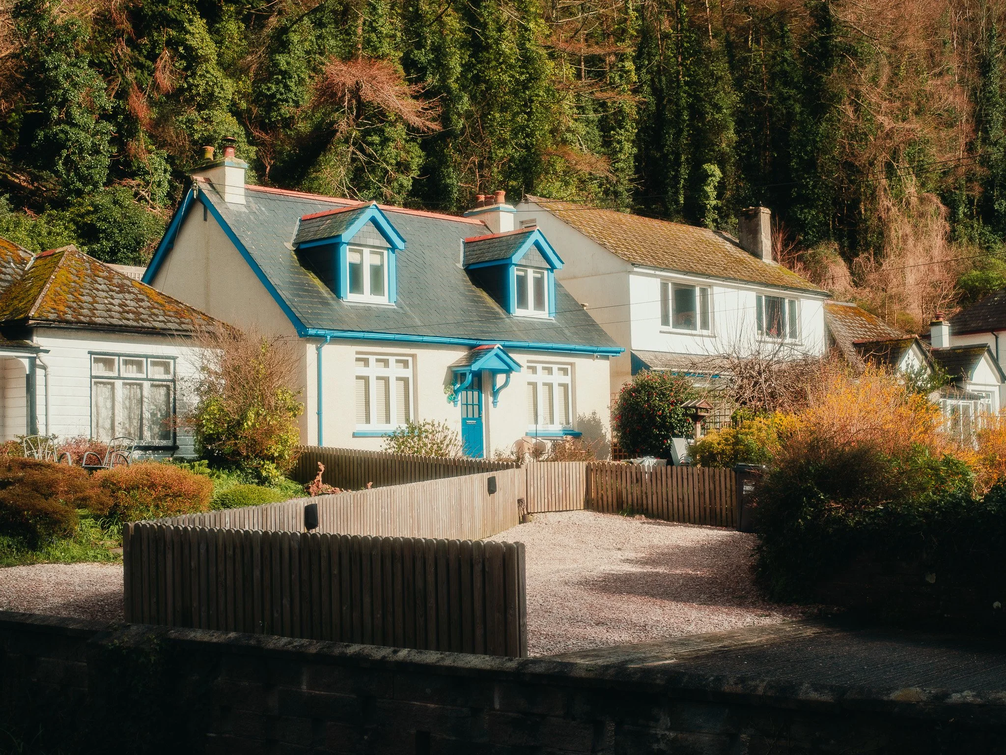 Houses with steep roofs near a wooded hillside with autumn trees, enclosed by a wooden fence.