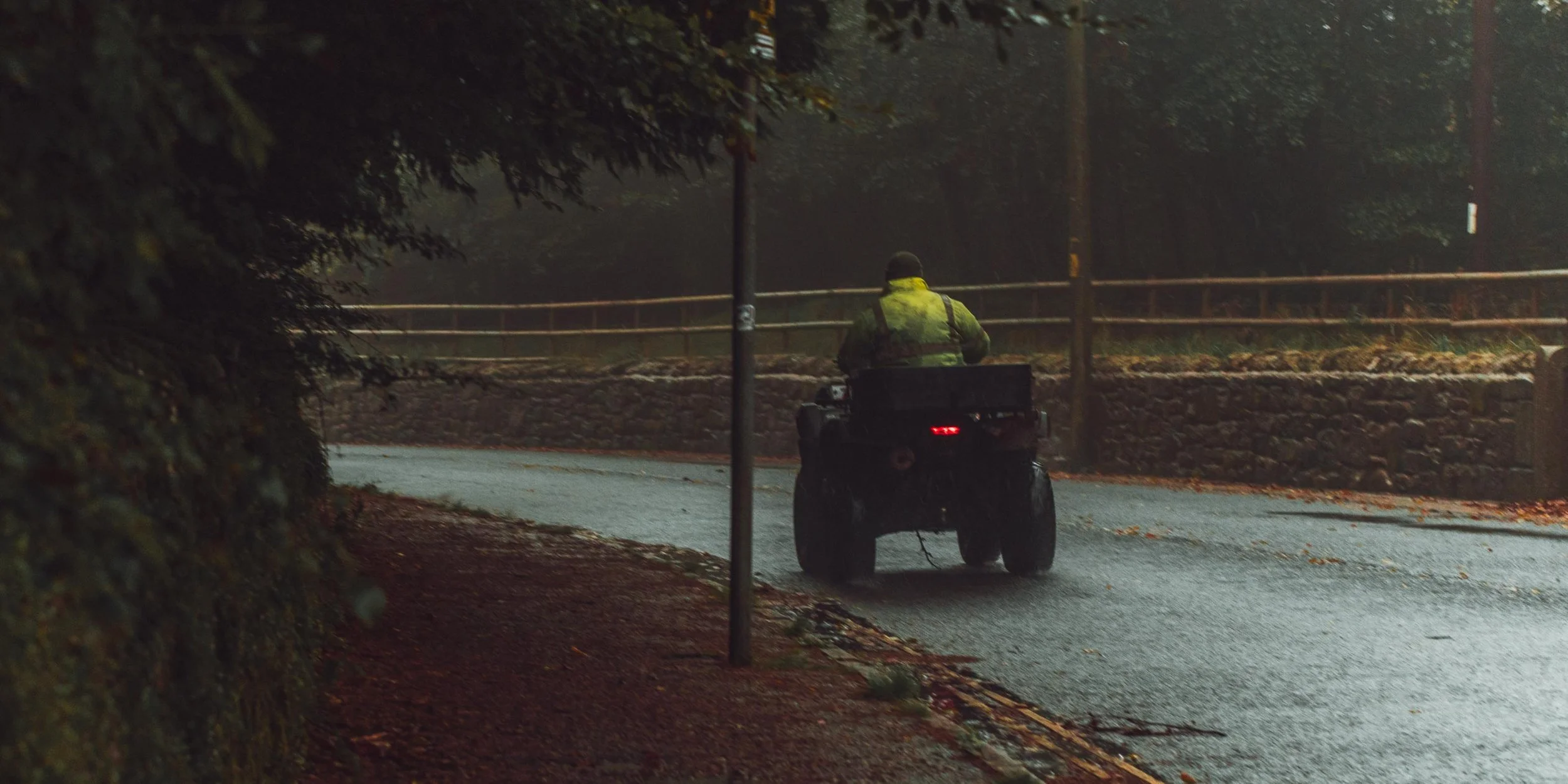 A person wearing a yellow safety vest riding a quad bike down a wet street at night, with trees and a fence along the sidewalk.