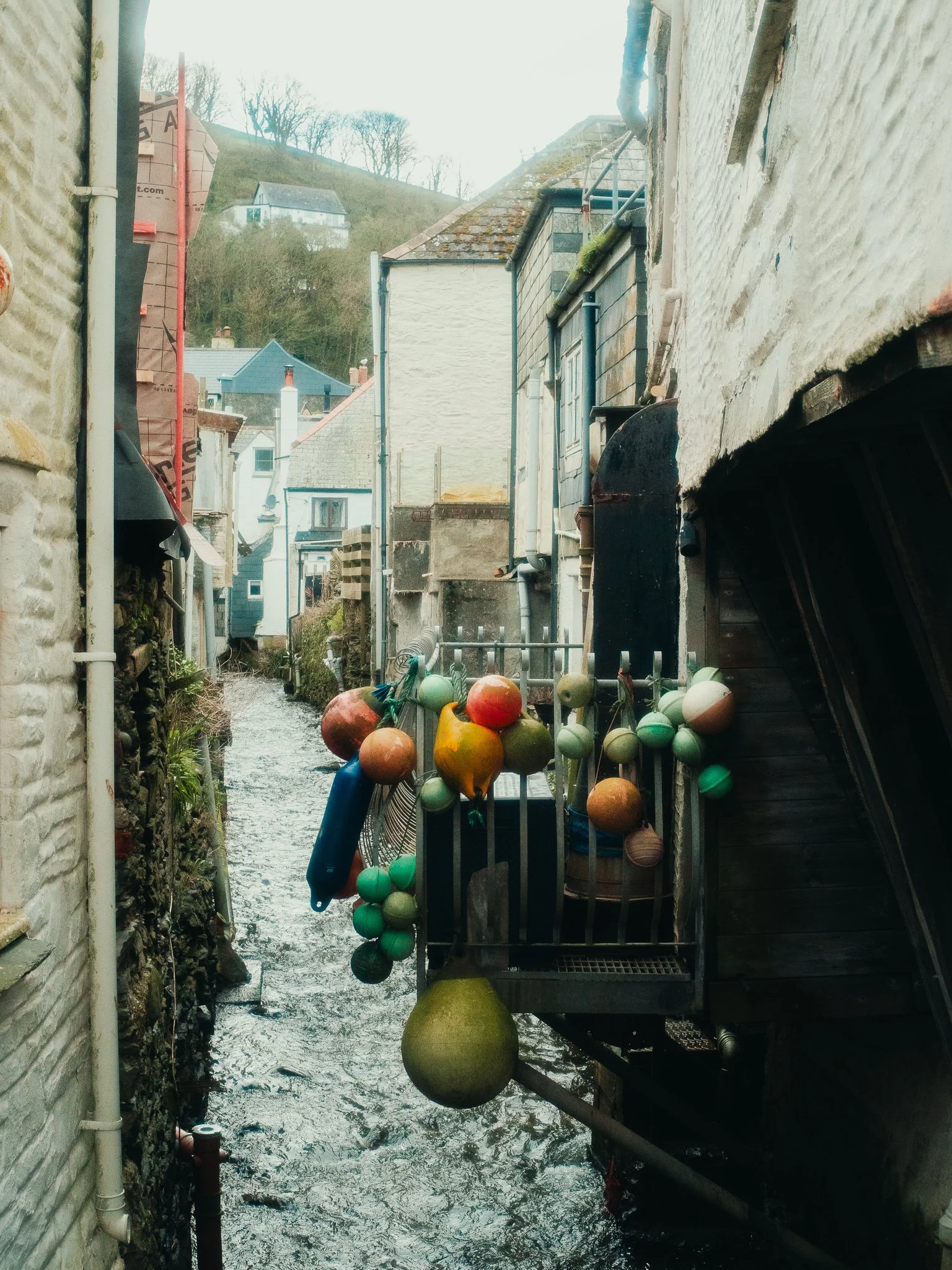 Narrow alley between two buildings with water flowing underneath, docked buoys hanging on a metal fence, and houses on a hillside in the background.