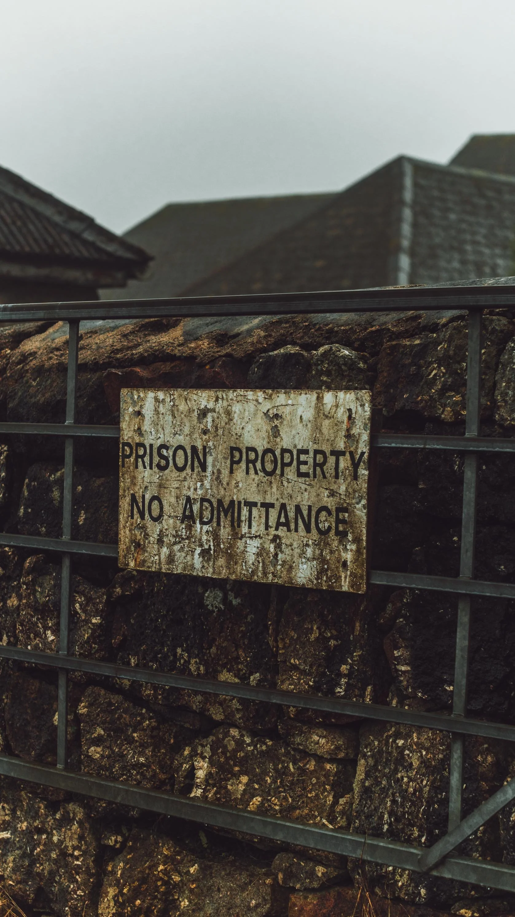 A rusty sign on a stone wall reads 'Prison Property No Admittance' and is attached to a metal fence with houses in the background.