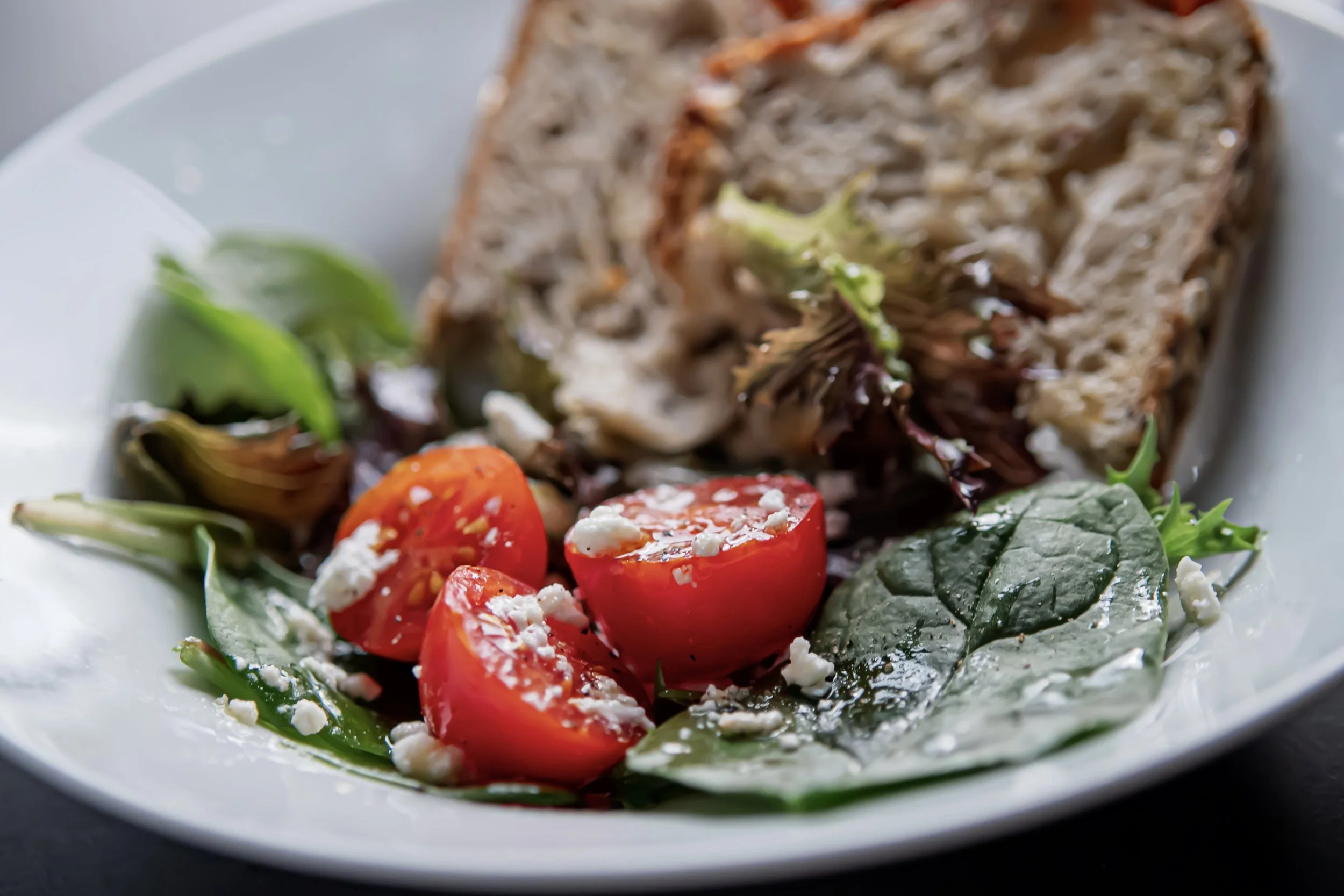 Salad with cherry tomatoes, spinach leaves, crumbled cheese, and a slice of bread on a white plate.