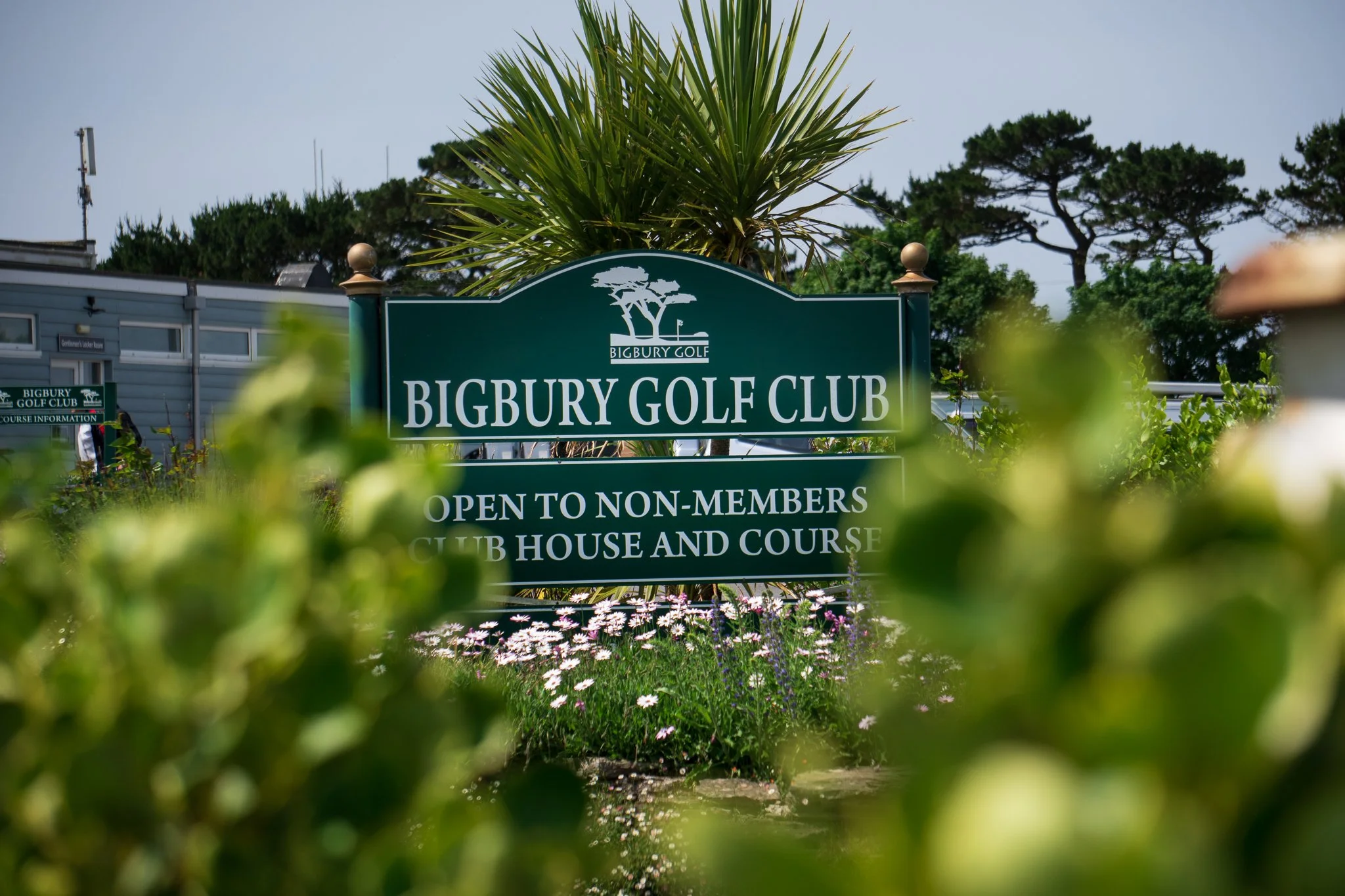 Sign for Bigbury Golf Club surrounded by green plants and flowers with trees in the background.