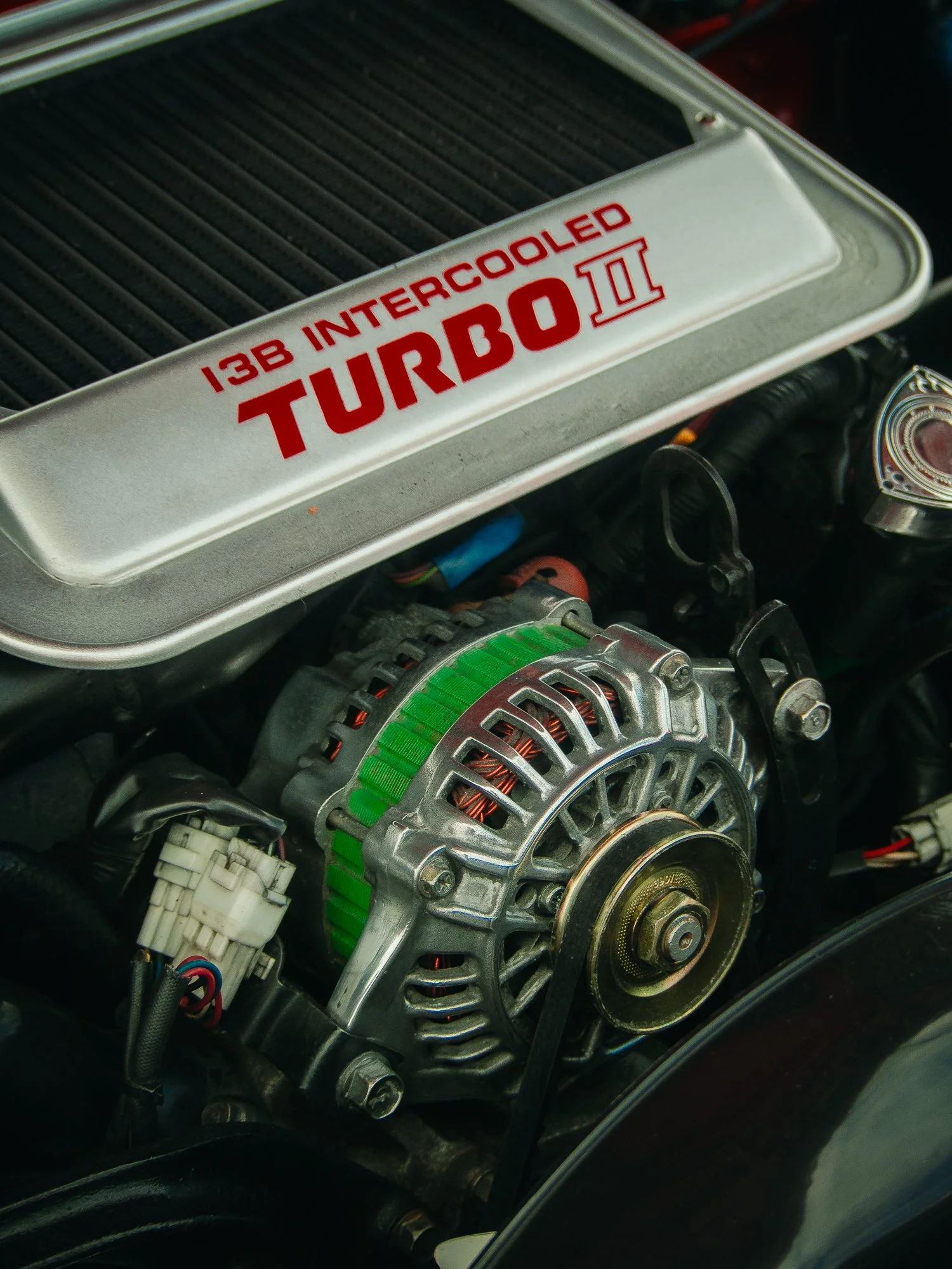 Close-up of a car engine with a silver supercharger labeled '13B Intercooled Turbo II' in red, with visible components including the alternator and wiring.
