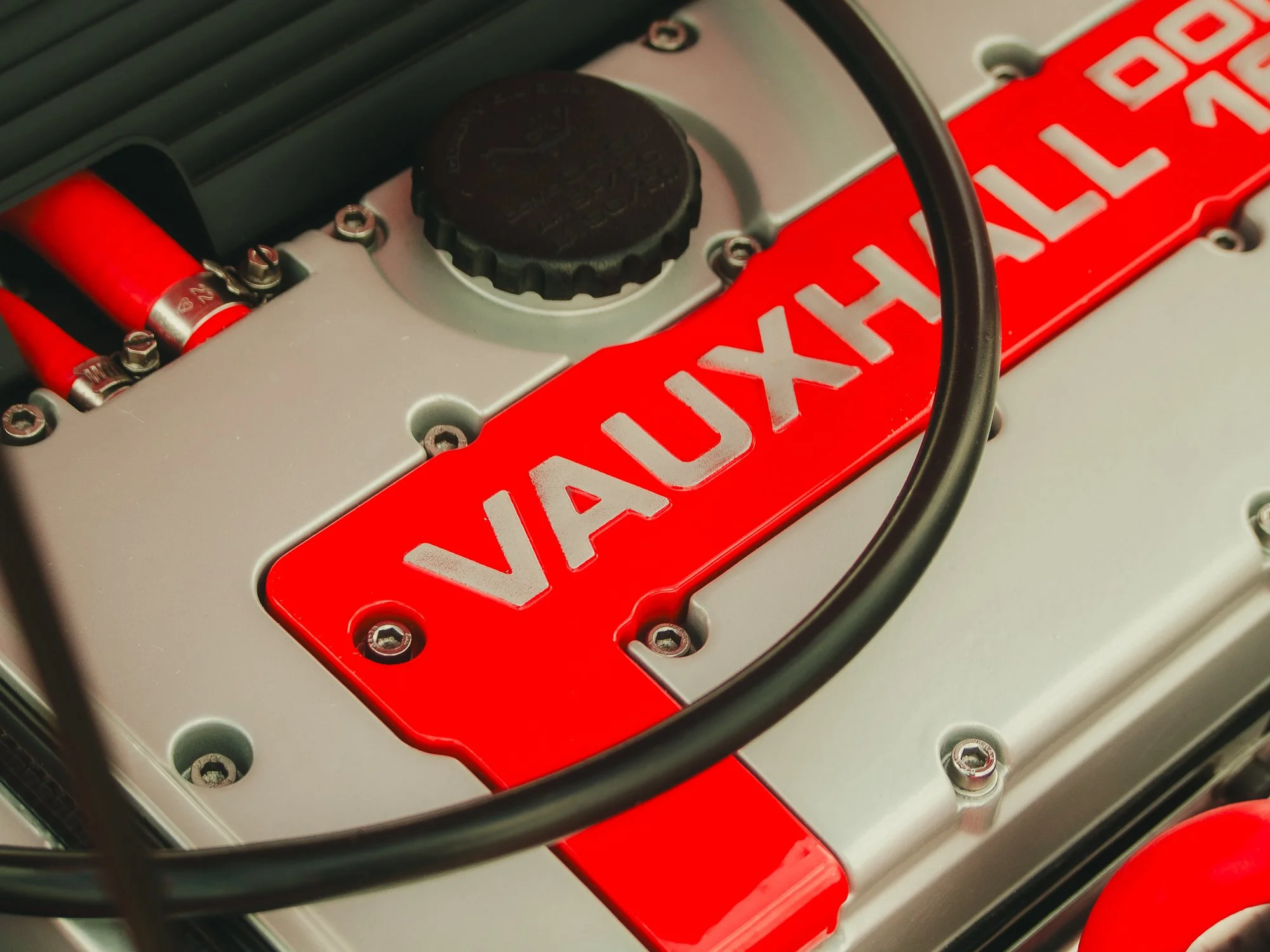 Close-up of a race car engine with a red valve cover and aluminum components. A black oil cap is visible, along with various bolts and hoses, including a red hose with a silver clamp. The engine has a prominent red and white "VALVE" lettering on the 
