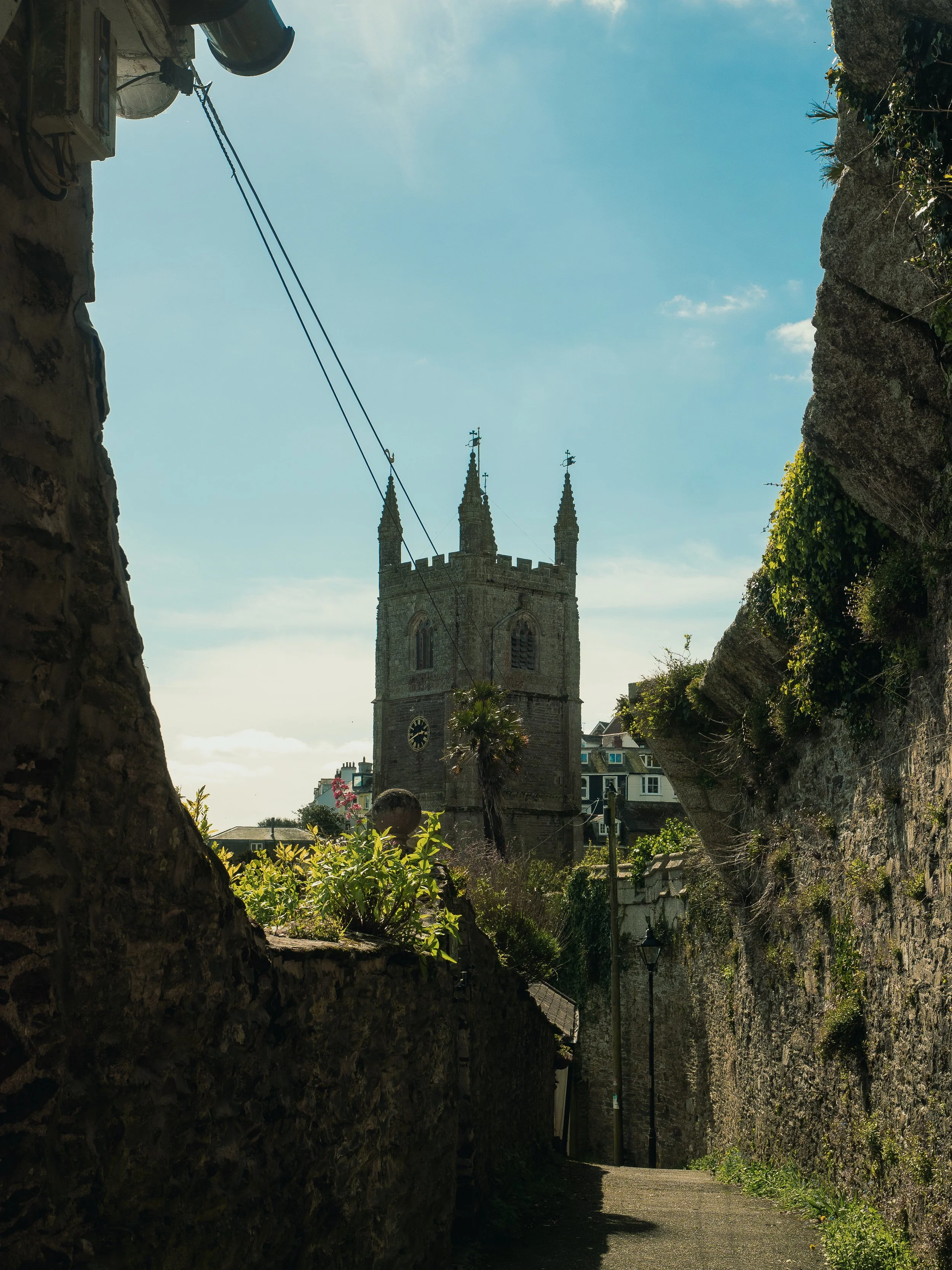 A narrow alleyway between stone walls with a view of a historic church tower with gothic architecture and clock, under a bright sky, with some greenery and houses in the background.