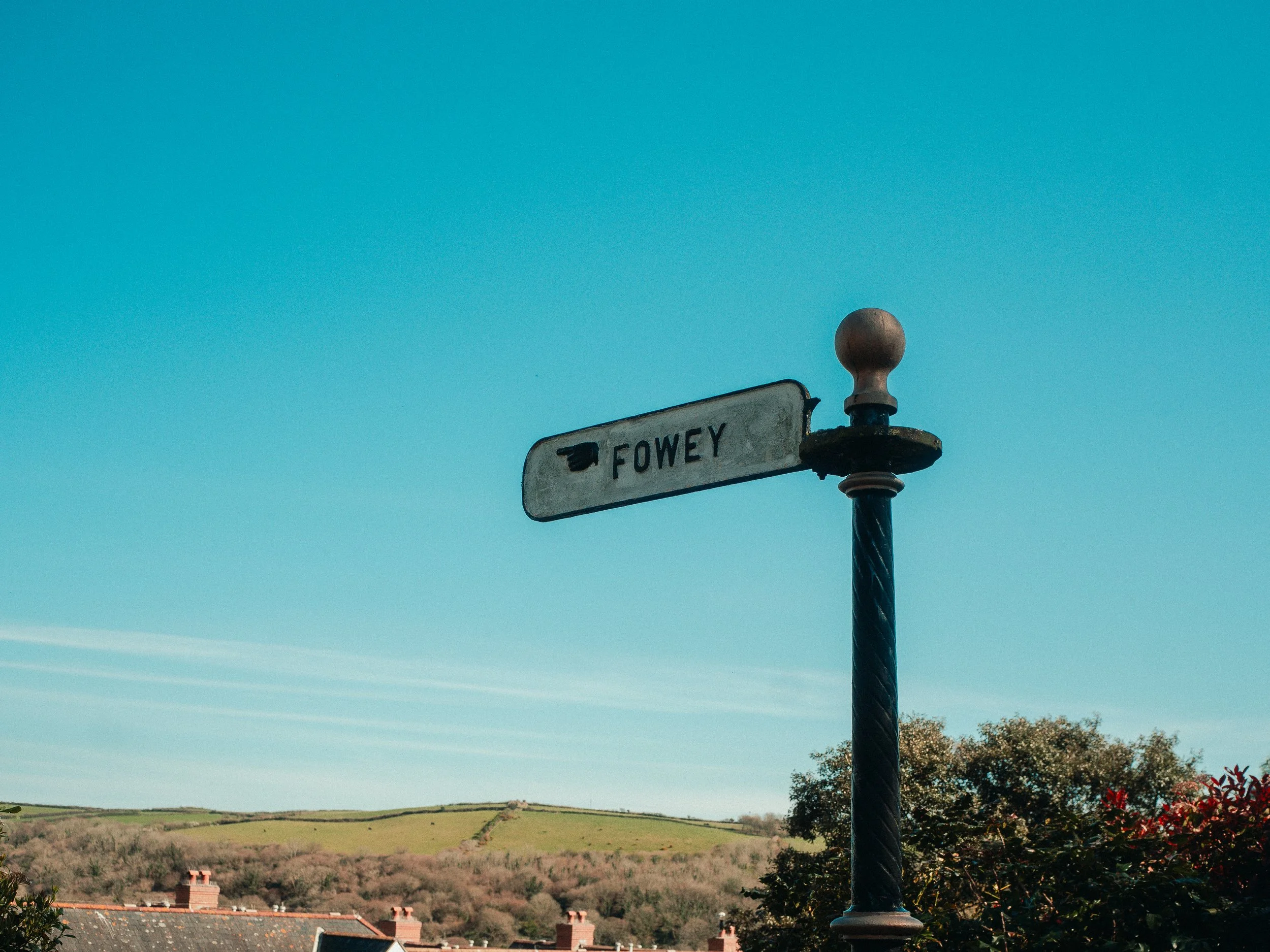 Street sign with the name 'Fowey' mounted on a decorative pole against a clear blue sky. In the background, there are rooftops of houses and rolling green hills.
