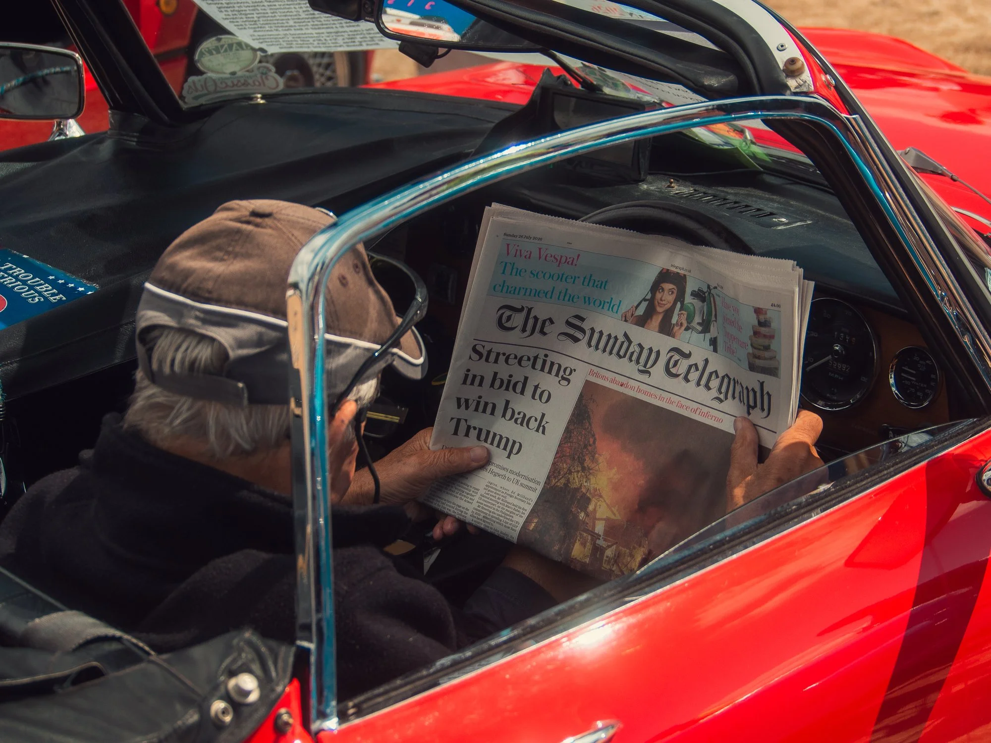 A person with gray hair reading a newspaper inside a red vintage car.
