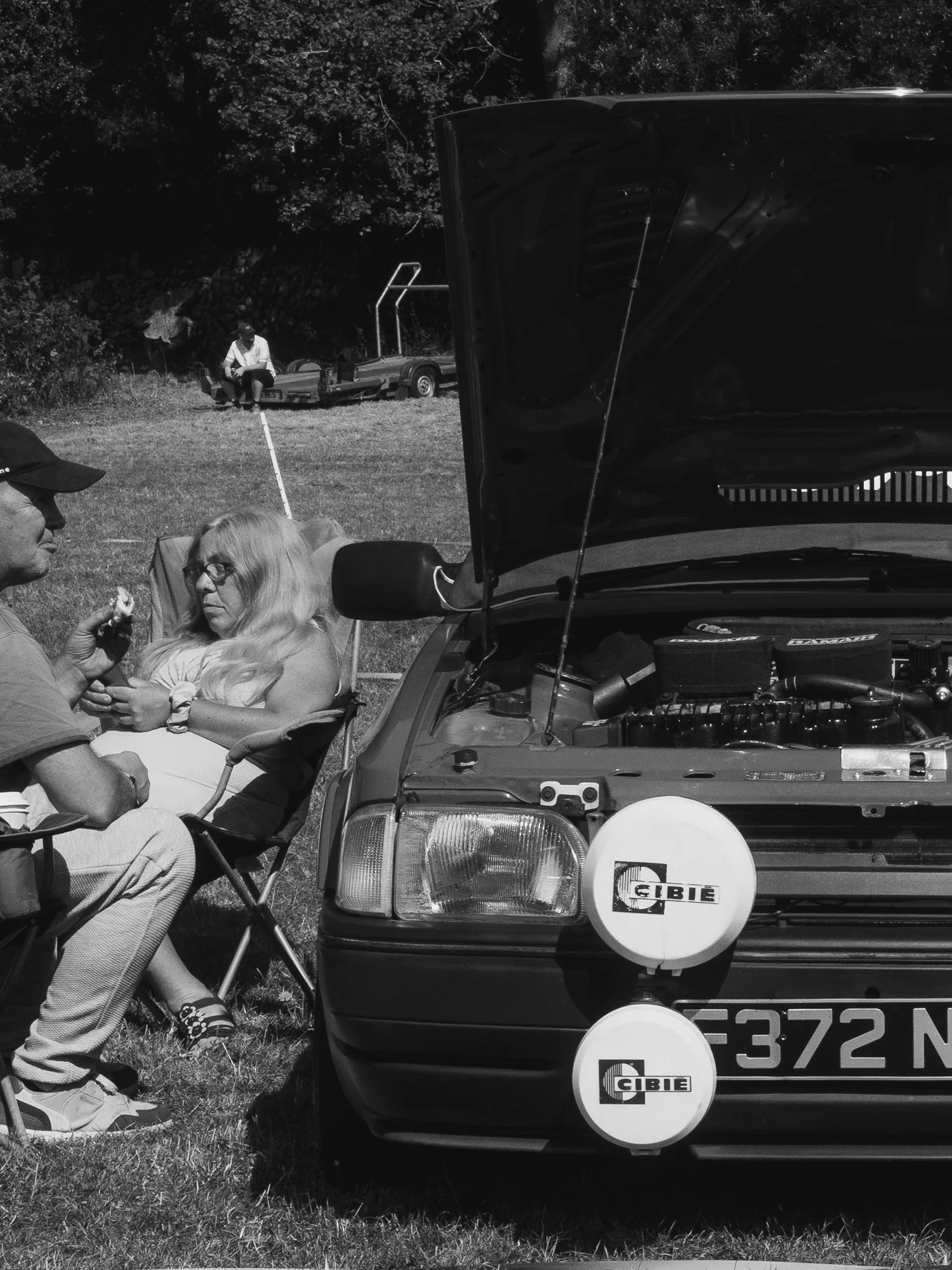Black and white photo of a group of people outdoors around a car with its hood open, with a man sitting on a trailer behind a man sitting on a folding chair, a woman sitting on a camping chair, and a man holding a small animal, possibly a dog, while 