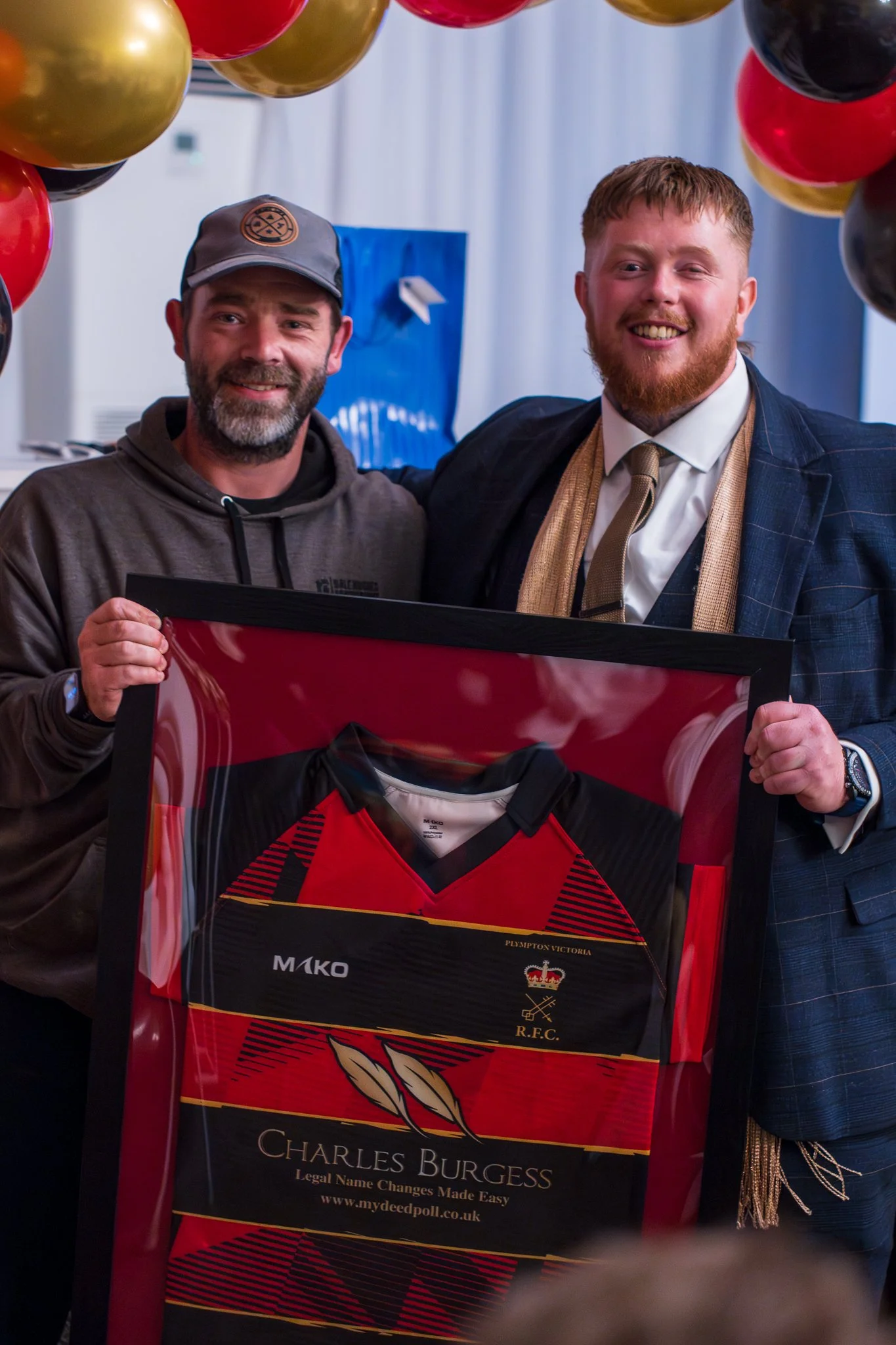 Two smiling men posing together at a celebration, one in a casual hoodie and baseball cap, and the other in a suit, holding a framed rugby jersey. Balloons are hanging above them, and a blue gift bag is in the background.