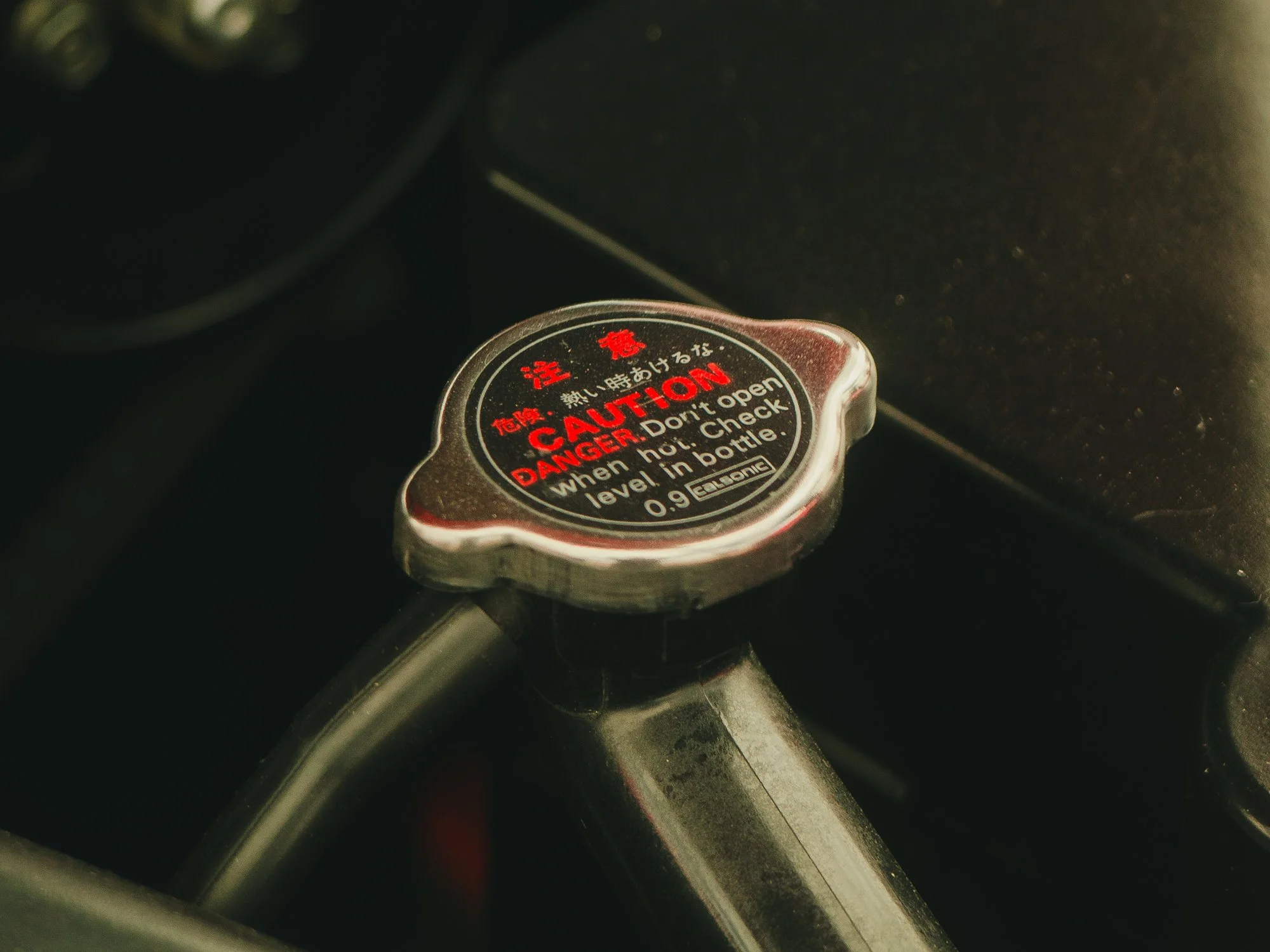 Close-up of a warning cap on a vehicle's radiator, with black and red warning text, partly visible brake or other component in the background.