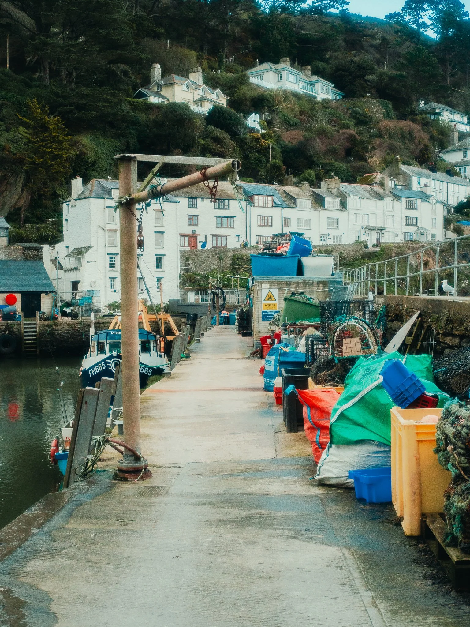 A harbor dock with various colorful bags and containers, a small boat docked on the water, and a hillside with houses and greenery in the background.